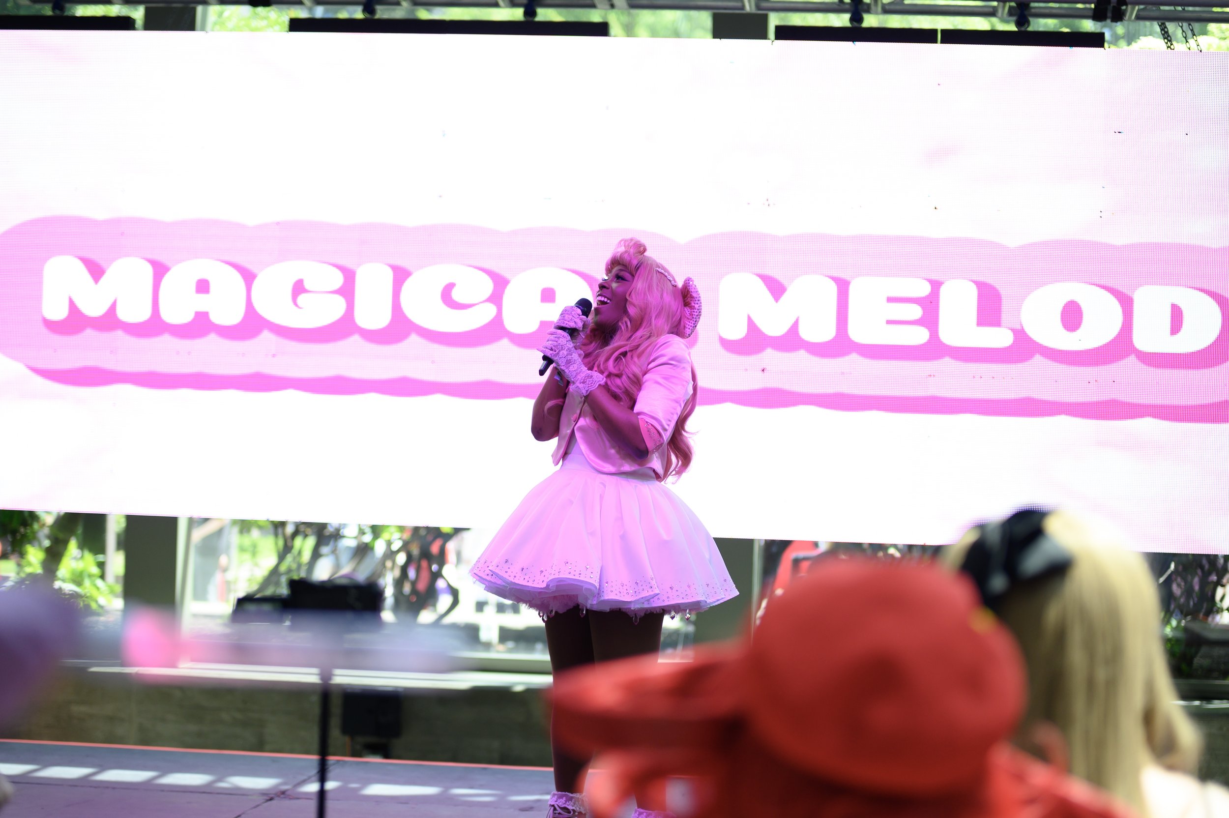 A performer dressed in a pink tutu, with long pink hair and fingerless lace gloves, singing into a microphone on stage. Behind her is a large screen displaying the words 'MAGICE AND MELOD' in pink and white. Audience members are visible in the foregr