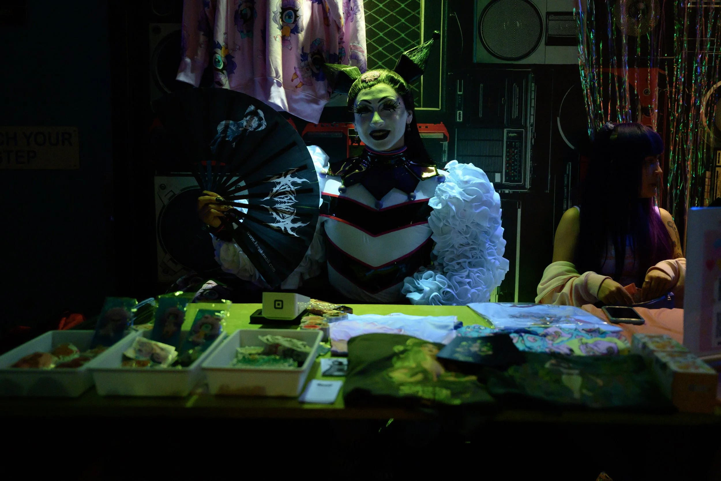 A person dressed in a flamboyant costume with a painted face, holding a black fan, sitting behind a table with various items in a dark, colorful setting.