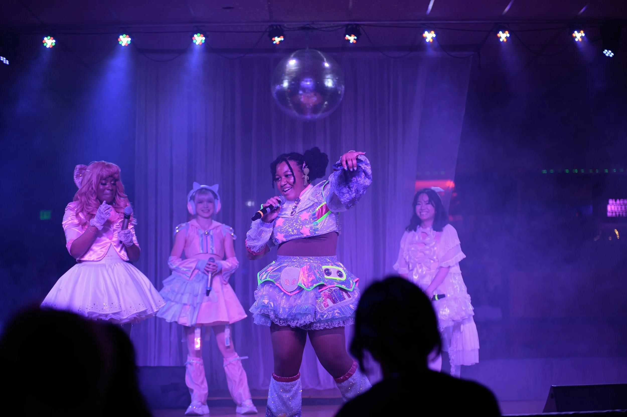 Four performers dressed in colorful, frilly costumes on a stage with purple lighting, engaged in a performance with microphones and dance moves, under a disco ball.
