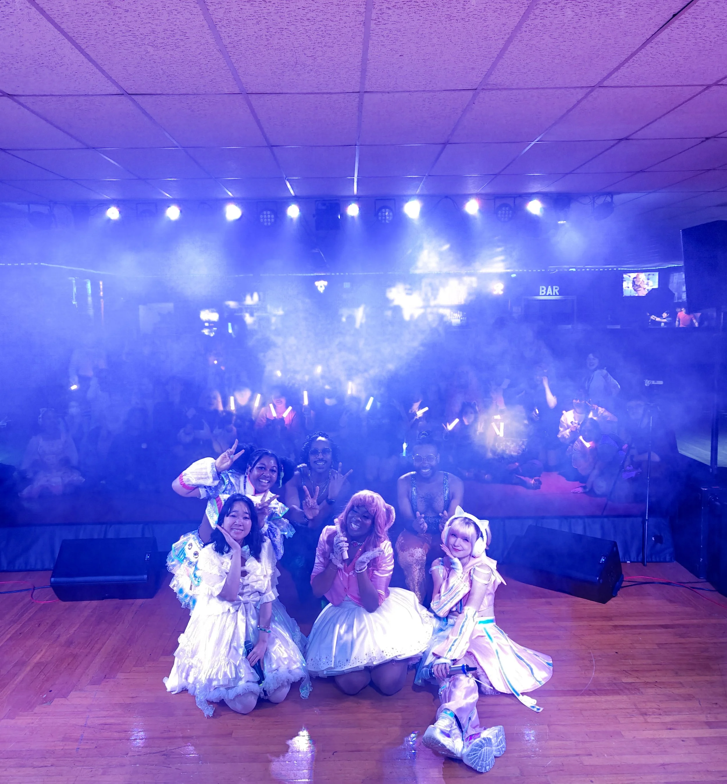 Group of young women dressed in colorful, playful costumes posing on stage with a foggy, lit-up dance floor and audience in the background.