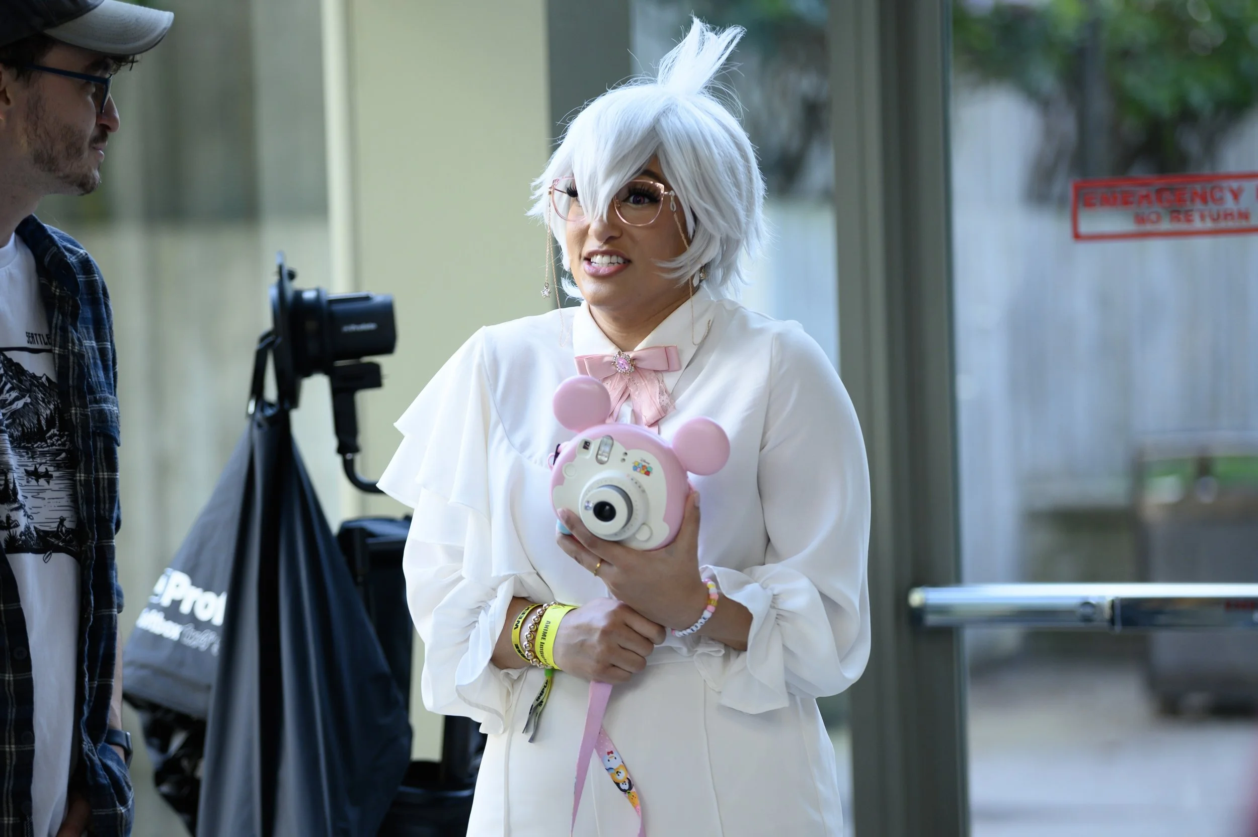 A woman with white hair and glasses wearing a white dress with a pink bow holds a pink camera shaped like a cartoon character.
