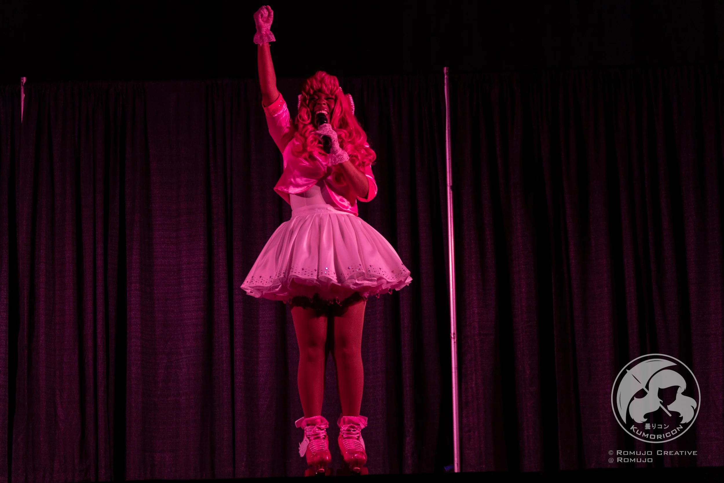 Performer dressed in pink with a tutu and pink shoes, singing on stage with a microphone, in front of black curtains.