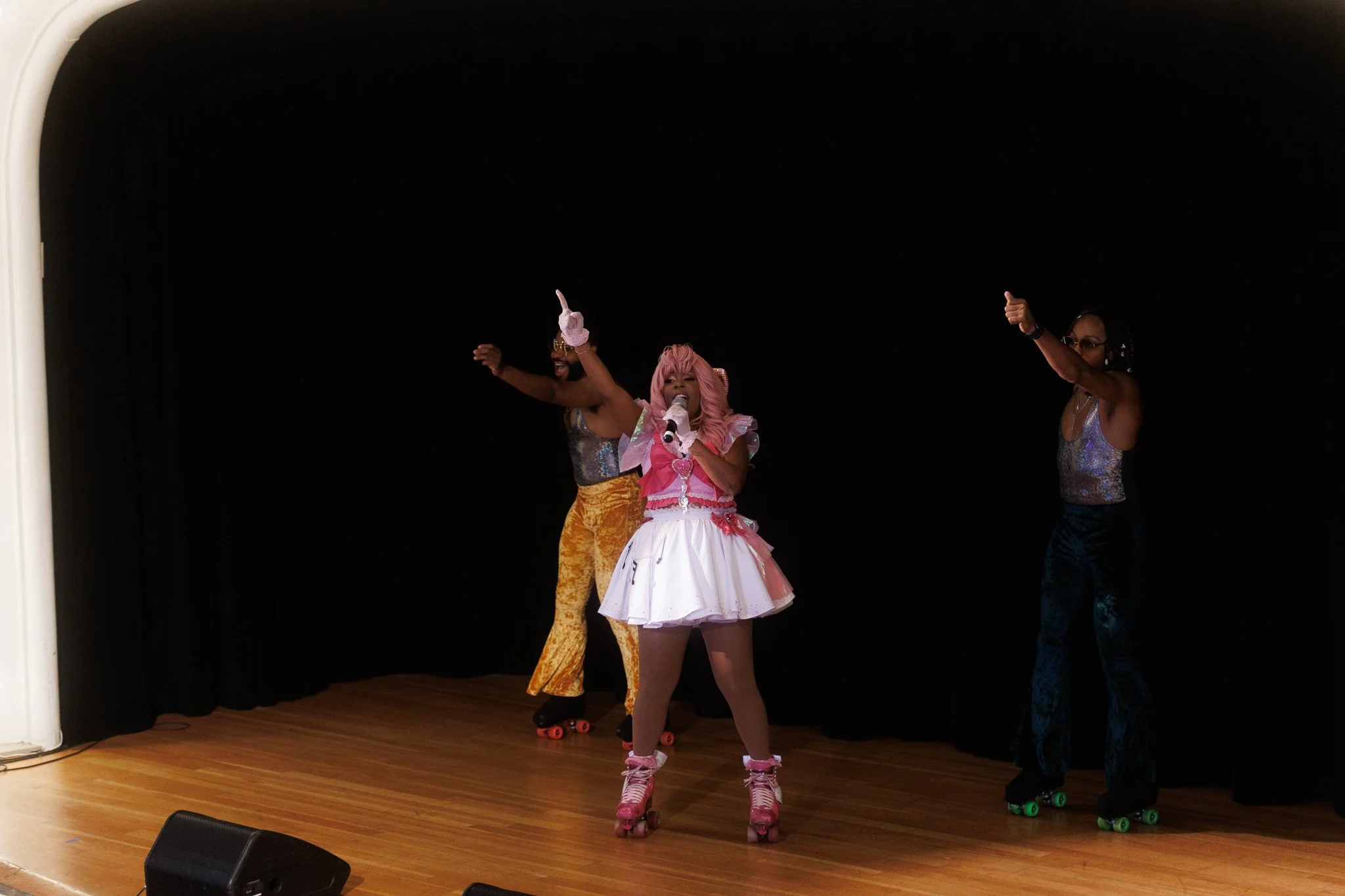 Three performers on stage roller-skating, with a woman dressed  in the center singing into a microphone. The woman in the center has pink hair and a pink and white tutu-style skirt, and the two performers on either side are wearing shiny tops and col