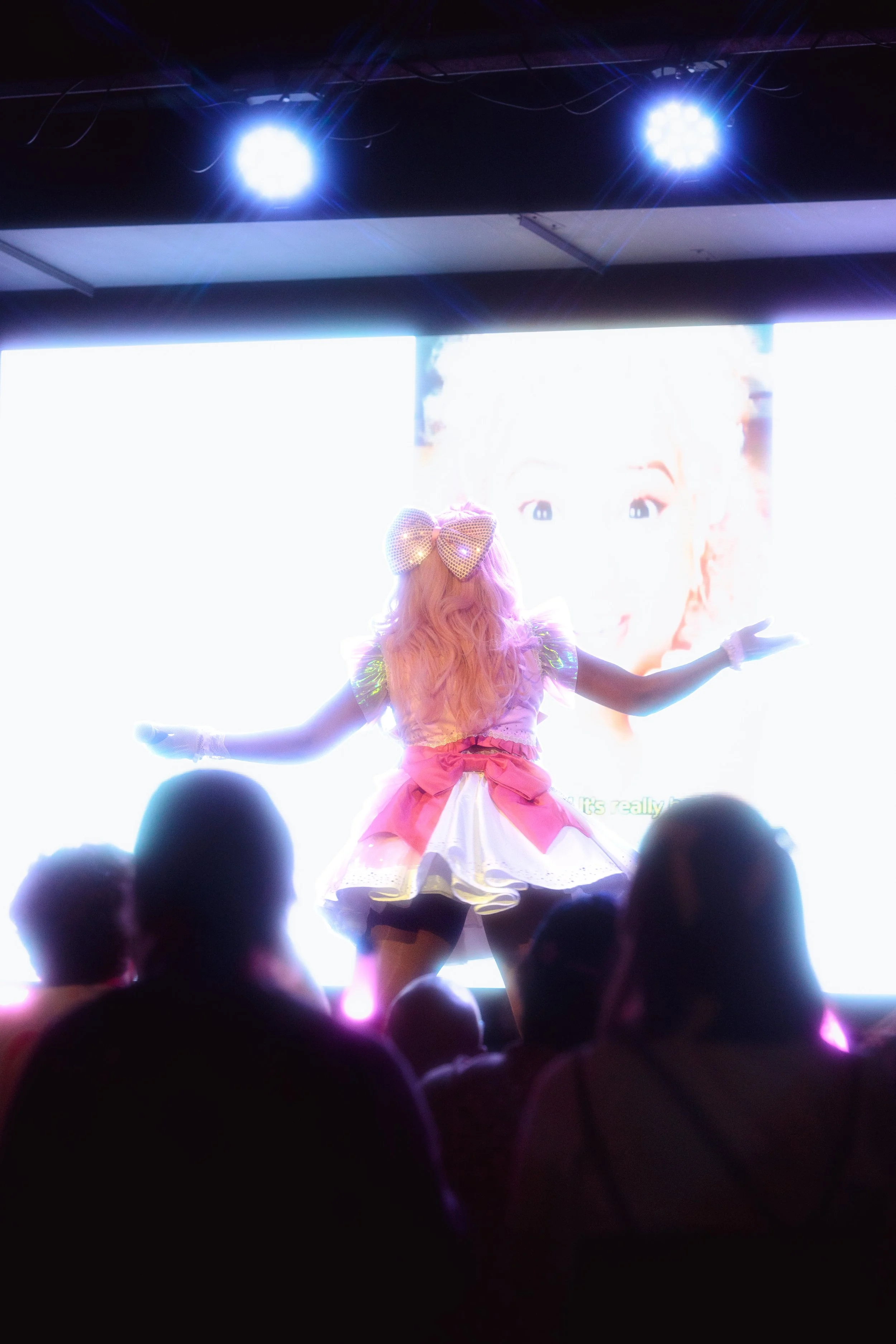 A performer dressed in colorful, anime-inspired costume with pink hair and large bow, dancing on stage in front of a bright LED screen with an anime character's face, with an audience watching.
