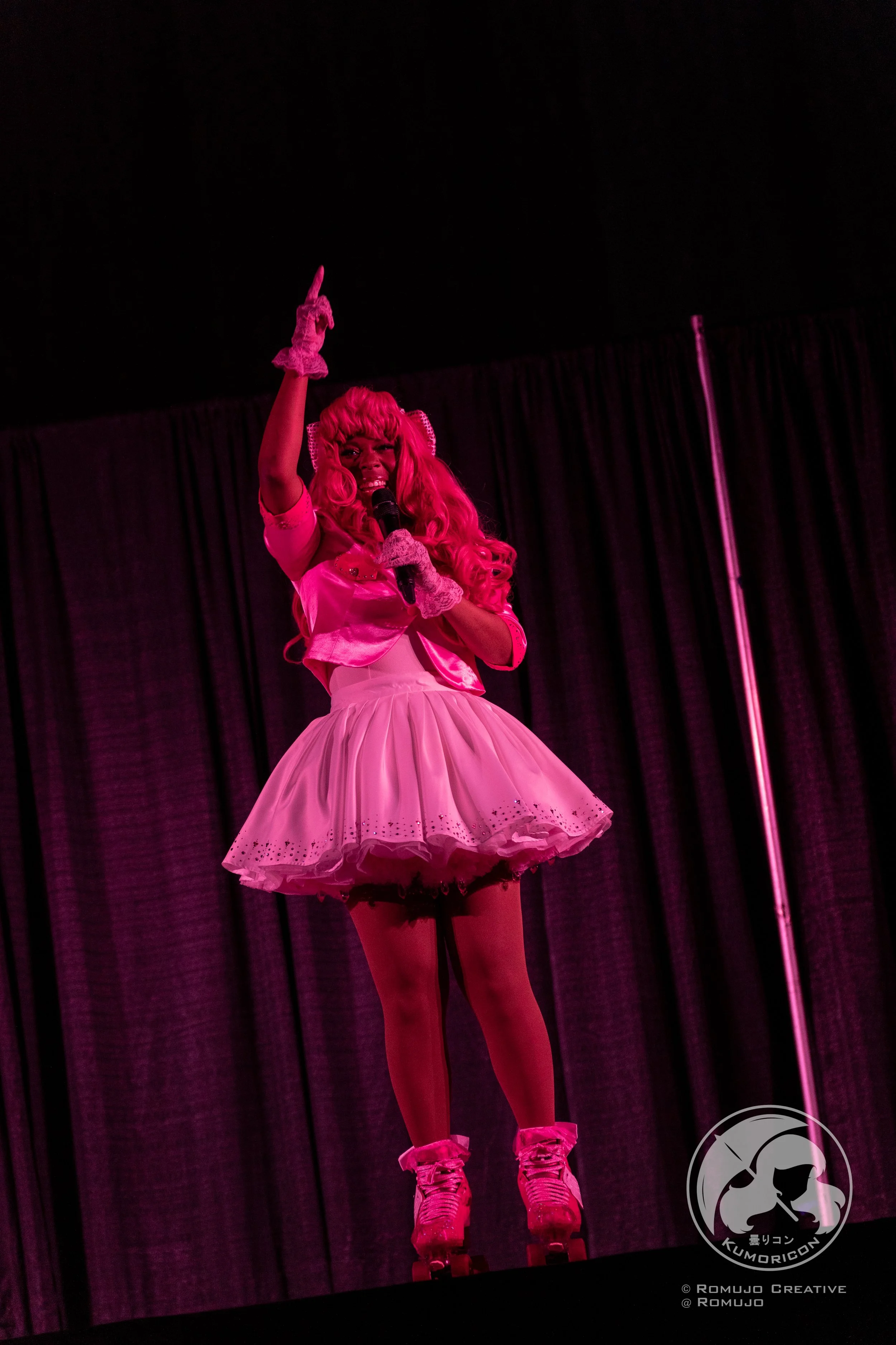 Performer dressed in bright pink cosplay costume with a voluminous skirt, wig, and roller skates, holding a microphone on stage with dark curtains in the background.