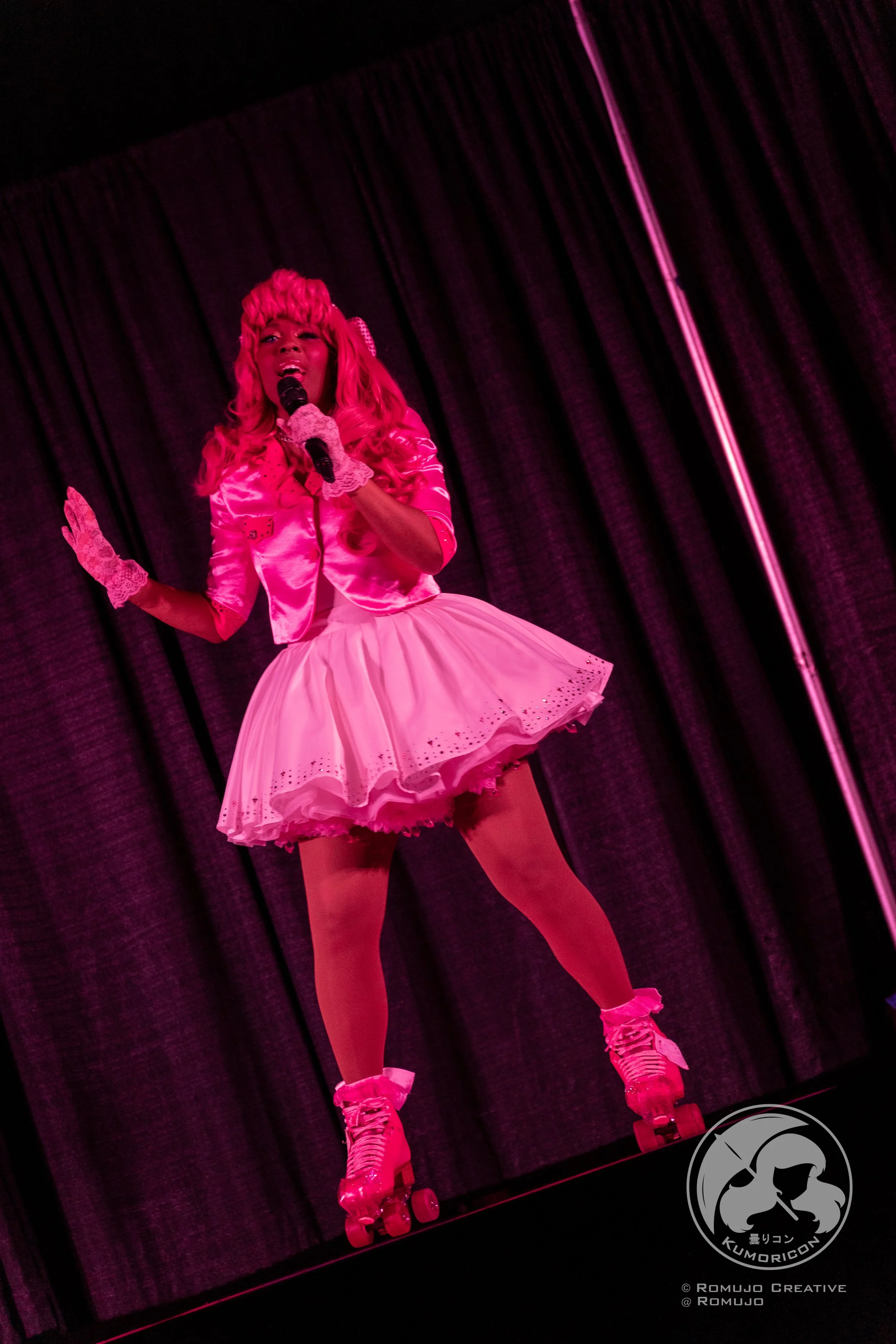 A woman performing on stage in a pink satin jacket, puffy skirt, and roller skates, holding a microphone with a black curtain backdrop.