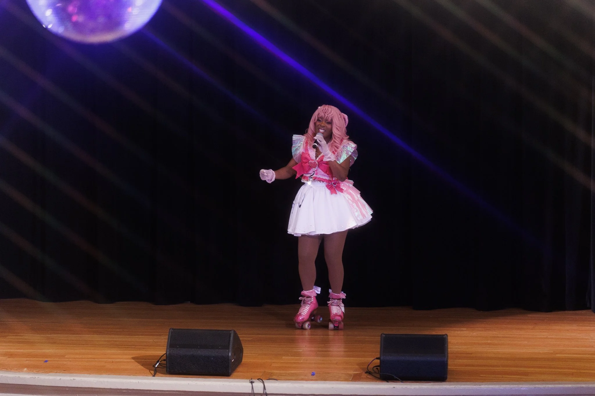 A woman dressed in pink and white cosplay outfit with roller skates performing on stage, holding a microphone, with a black curtain and a disco ball above.