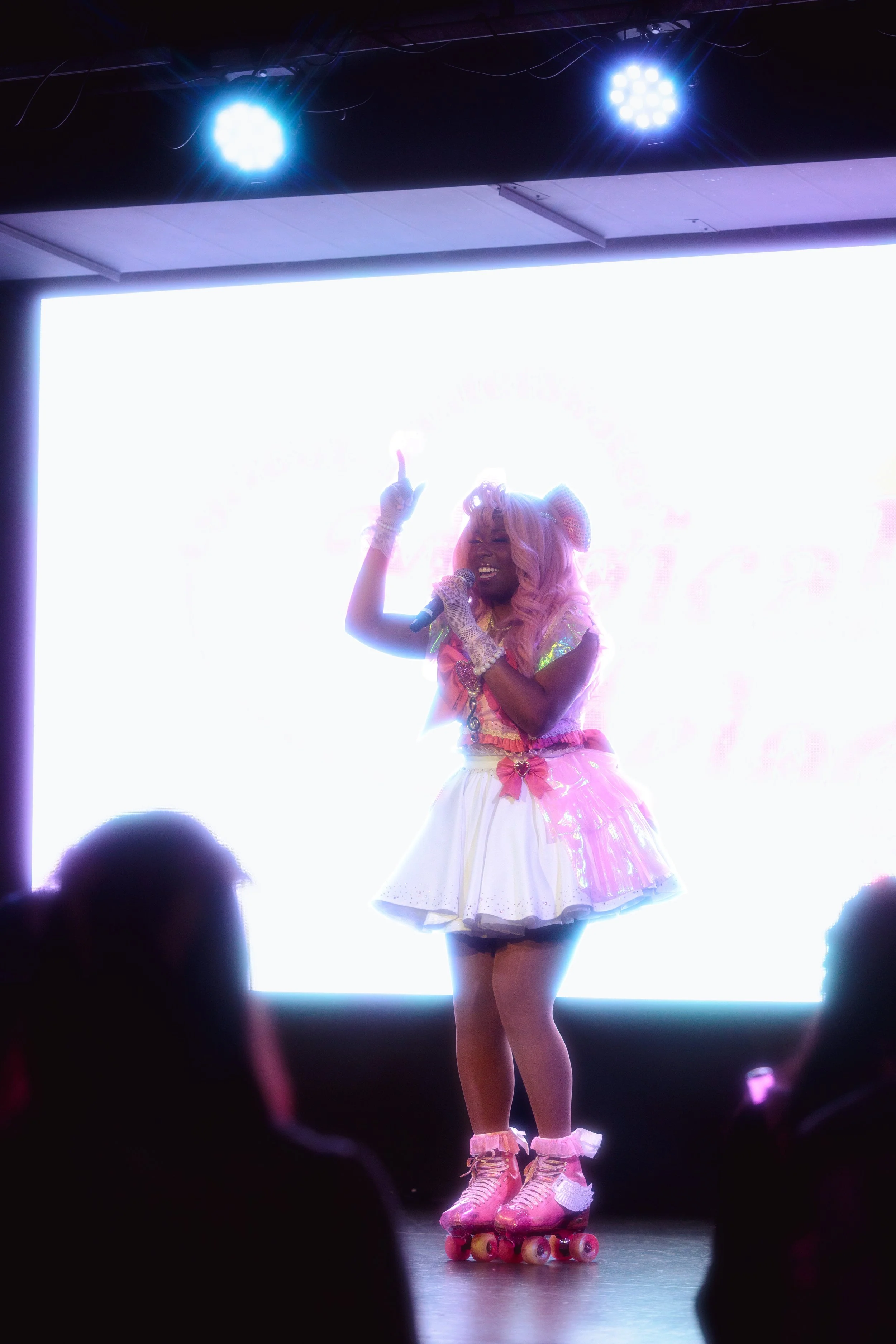 A woman in a colorful outfit and roller skates performing on stage with a microphone, lit by bright stage lights, in front of a large white screen.