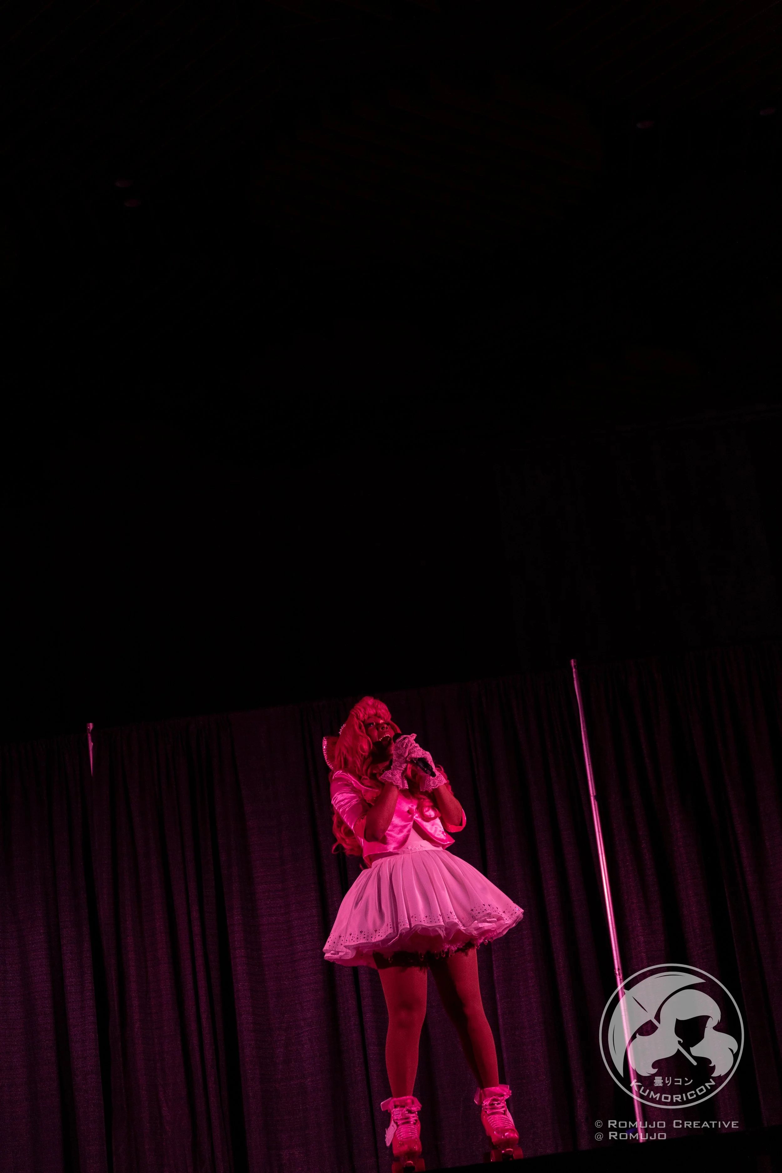 Performer in pink costume singing on stage with curtain background.