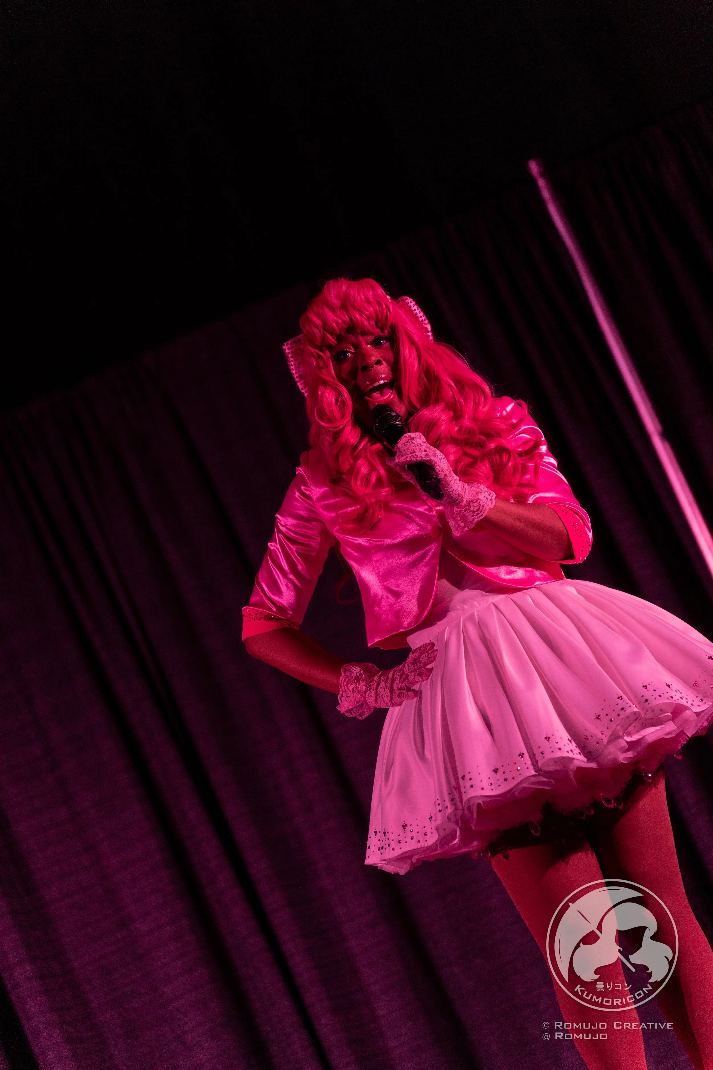 Performer dressed in pink, singing into a microphone on stage with dark curtains behind, in a pink-themed outfit with a tutu and gloves.
