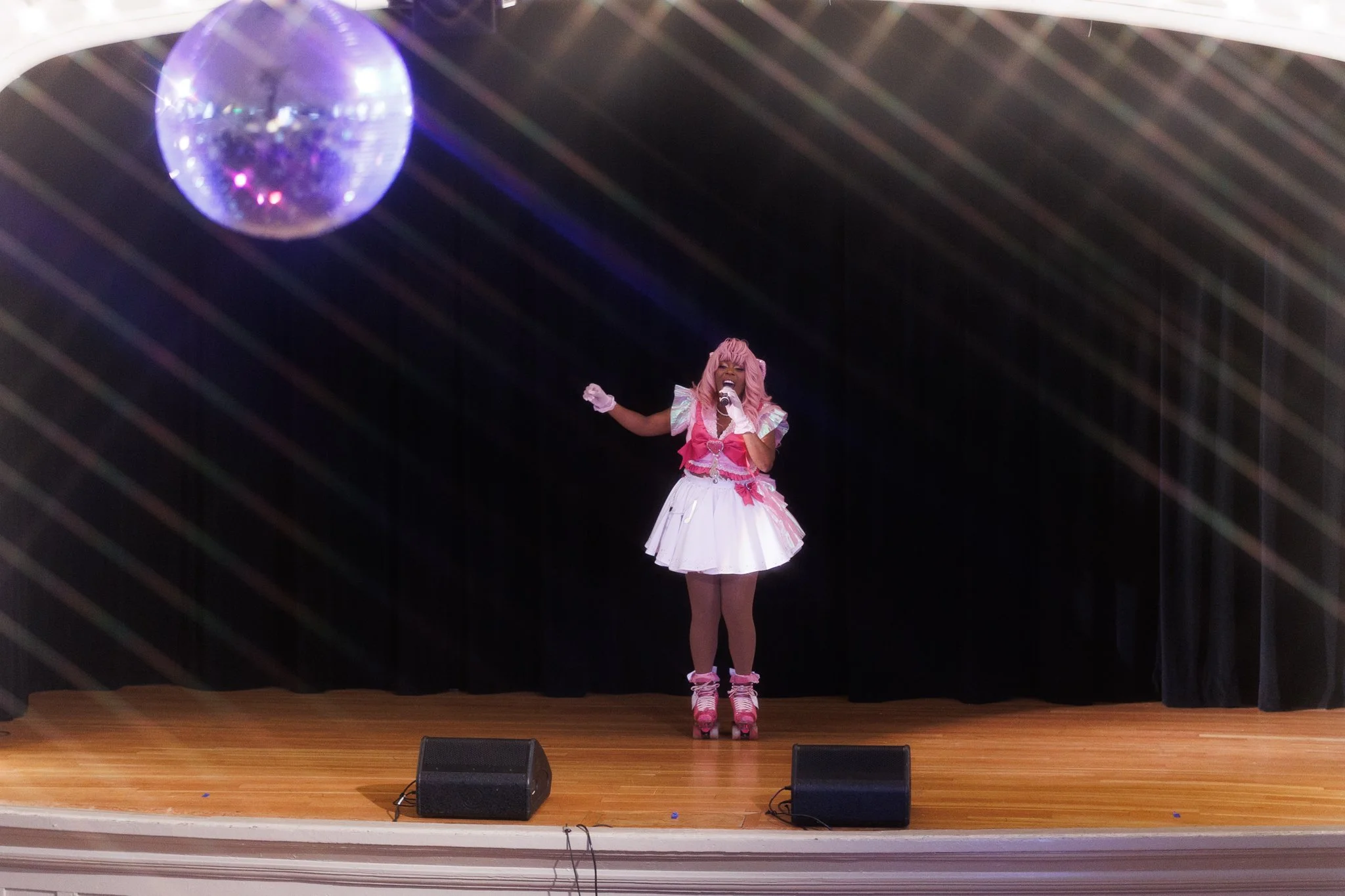 Performer in pink and white costume singing on stage with a disco ball overhead and black curtain backdrop.