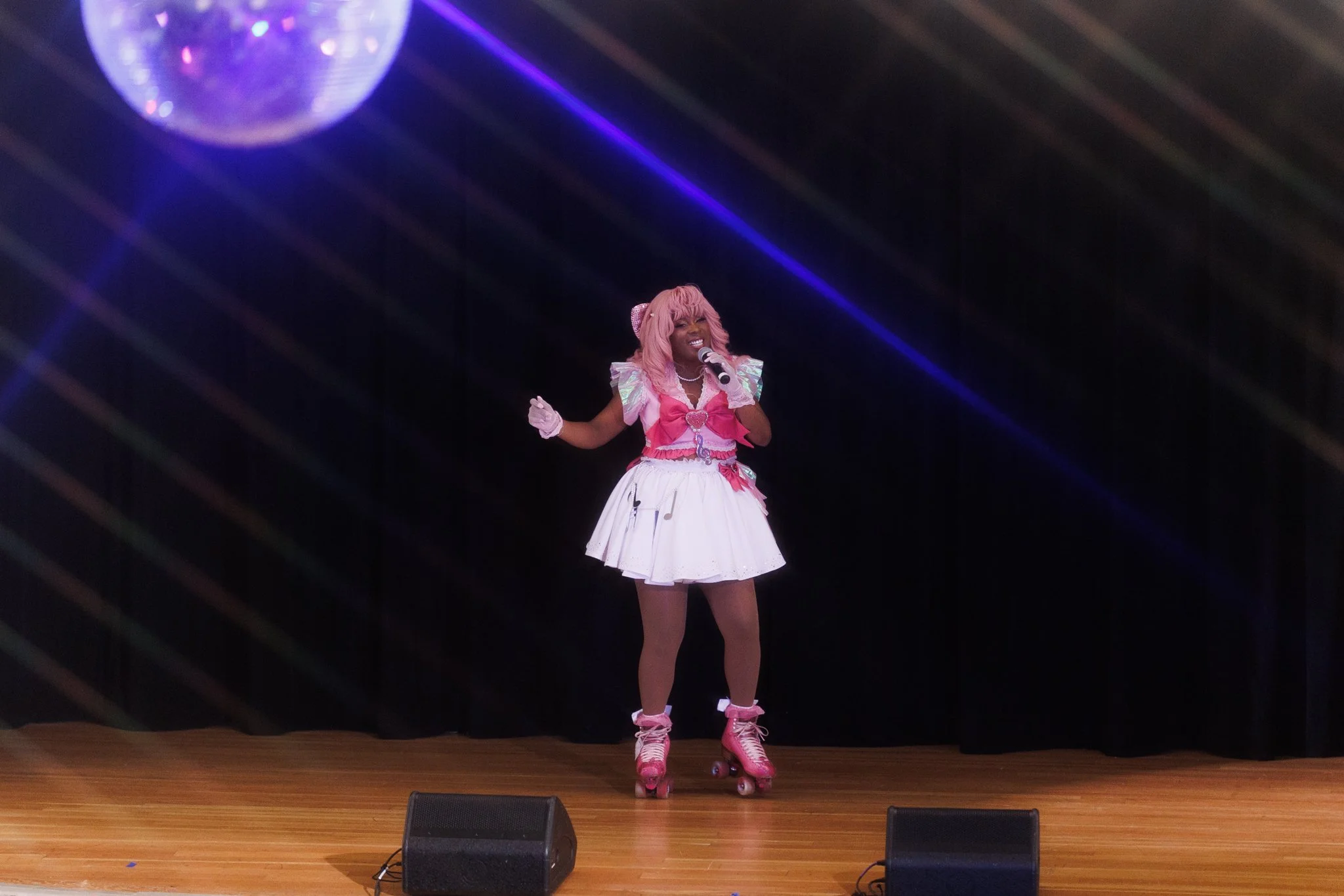 Performer at a stage wearing pink roller skates, a pink and white costume with ruffles, singing into a microphone, with a disco ball and stage lights.