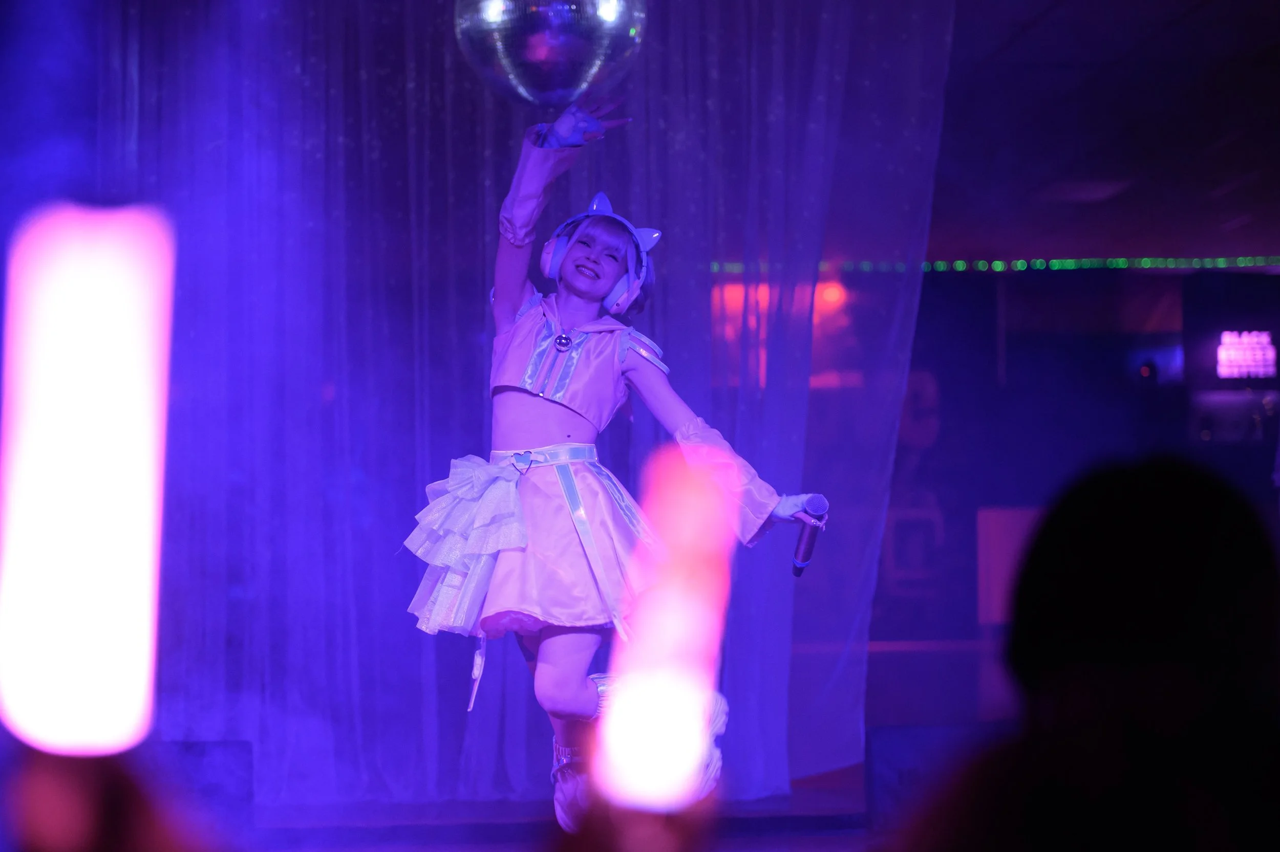 A performer on stage dressed in a pastel-colored costume with a skirt, top, and accessories, holding a microphone in one hand and reaching up towards a disco ball with the other, under colorful stage lighting.