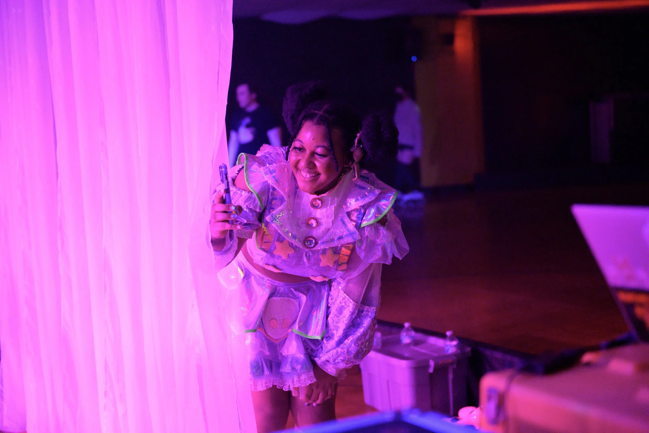 A woman with styled hair and a colorful, shiny costume smiling and taking a picture in front of a pink curtain at a party or event.