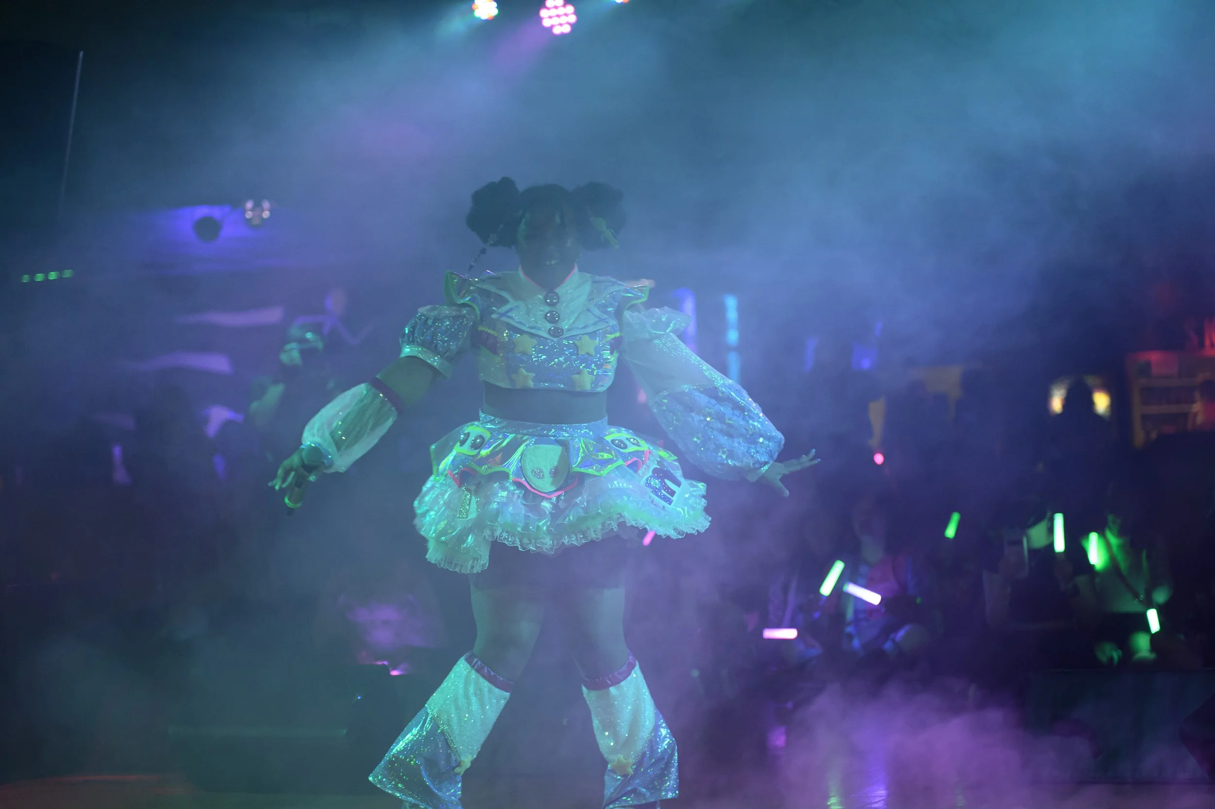 A person dressed in a colorful, sparkly costume with a patterned skirt, puffy sleeves, and leg warmers, performing on a stage with fog and rainbow-colored lights in a dark setting.