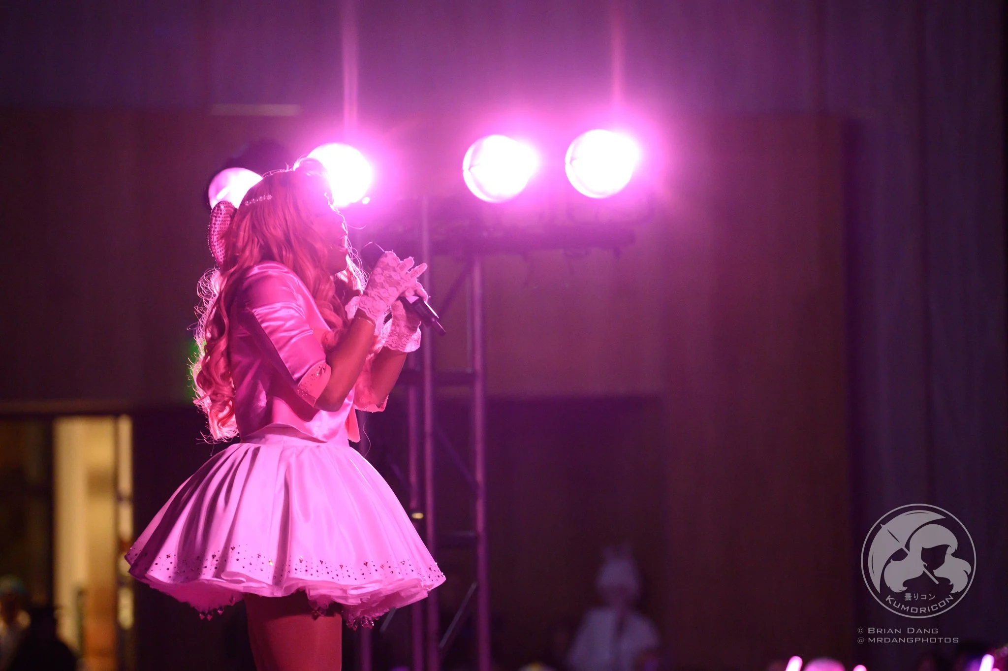Girl in pink dress with lace gloves singing into a microphone on stage under pink stage lights.
