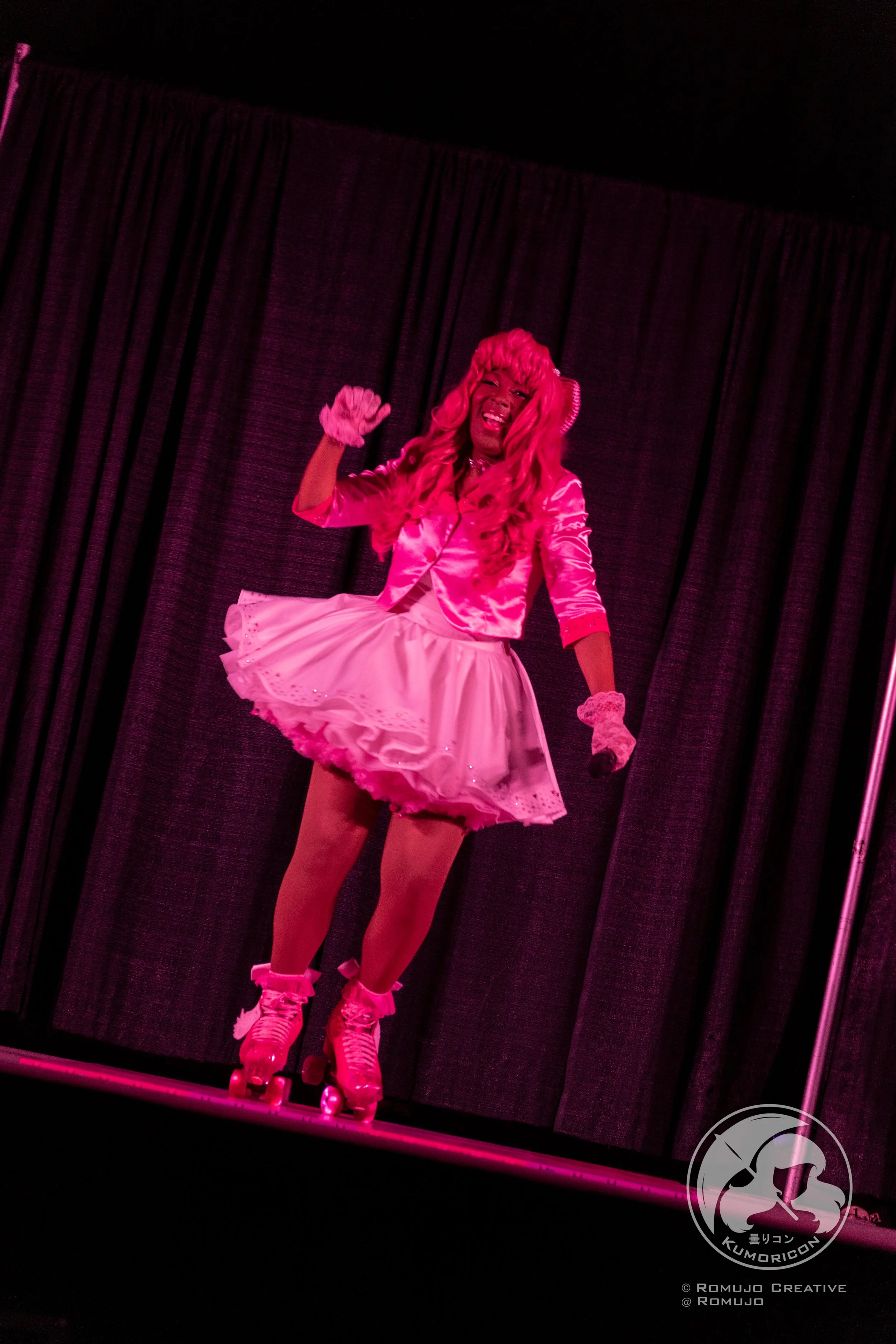 Performer in pink costume on stage with roller skates, pink skirt, and pink wig, holding a microphone, performing in front of dark curtains.