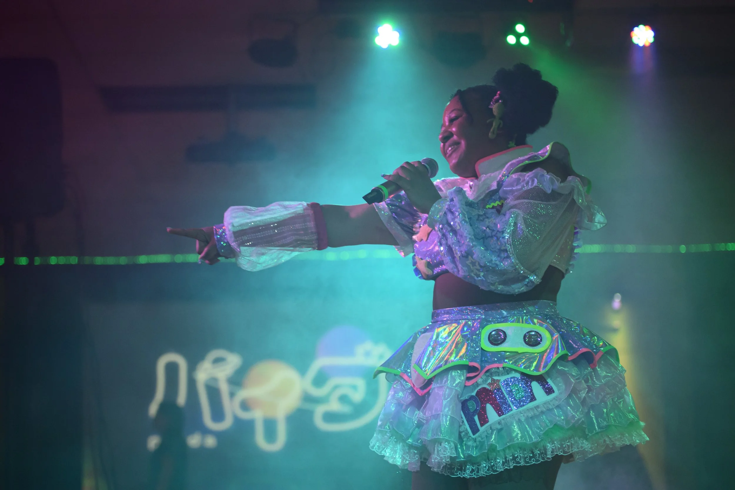 A young girl in a colorful, shiny, futuristic outfit holding a microphone on stage, pointing with her other hand, under green and blue stage lights with a digital background.