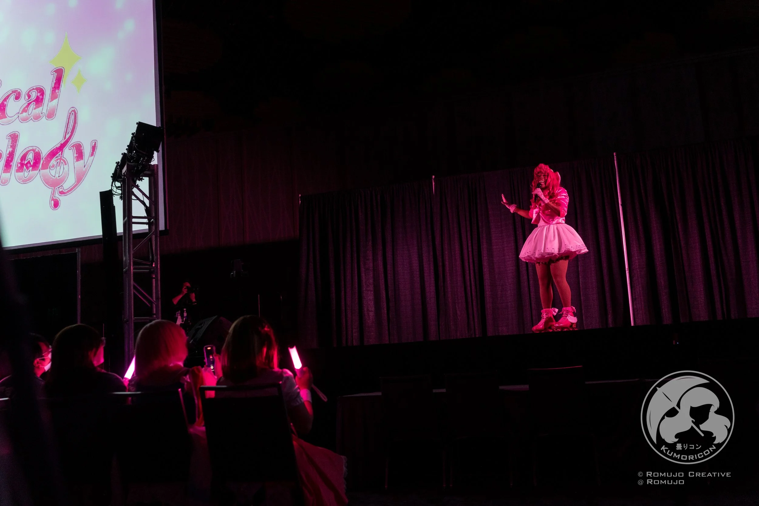 A performer dressed in pink, with pink roller skates, on stage at a convention or event, singing or speaking into a microphone. Audience members in the foreground are watching and recording.