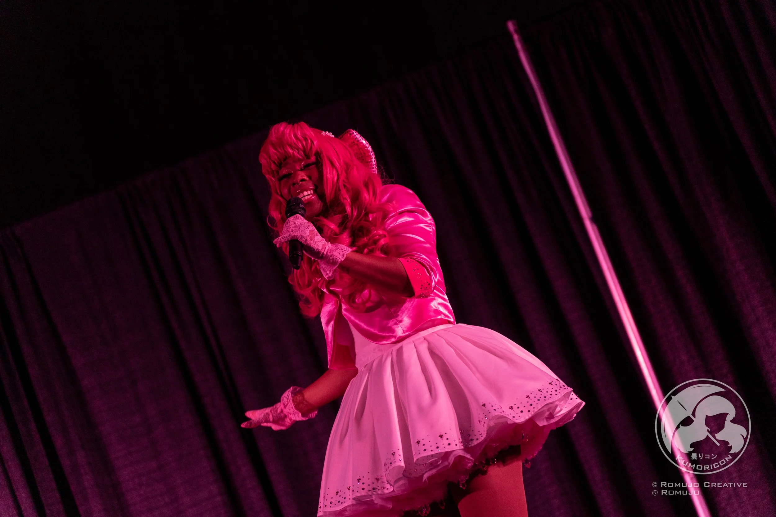 Performer dressed in pink with long curly hair, singing into a microphone on stage with a dark curtain backdrop.