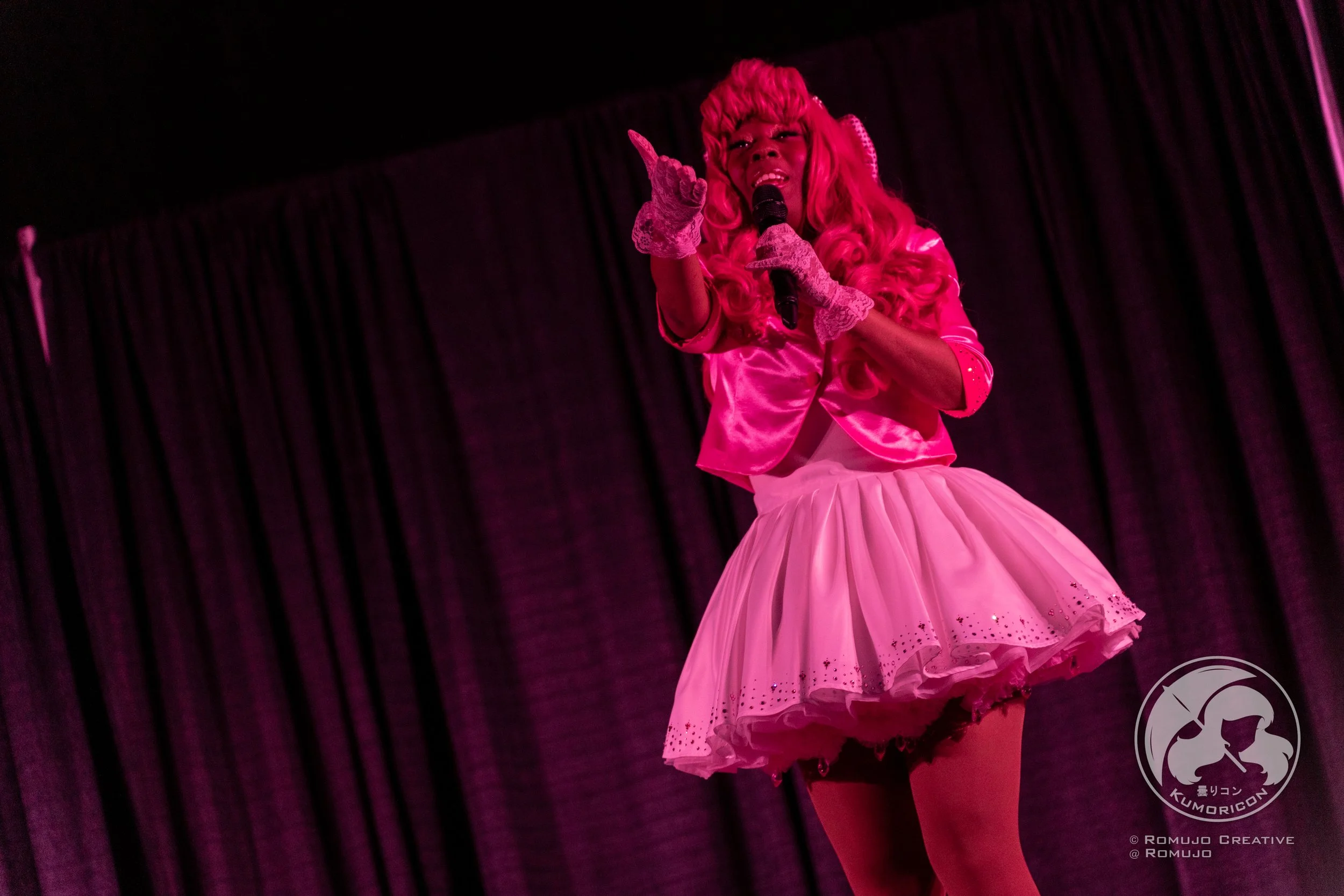 Performer dressed in pink satin top and pink tutu skirt singing on stage with a microphone, black curtain backdrop, and logo in lower right corner.