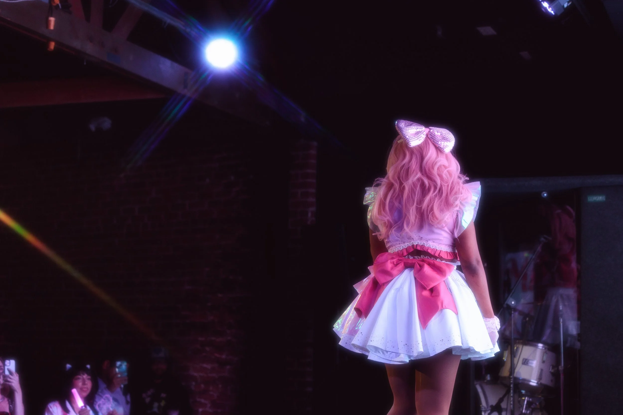 A performer with long pink curly hair dressed in a pink and white costume with a large pink bow at the back, standing on stage with a dark background and bright stage lights.