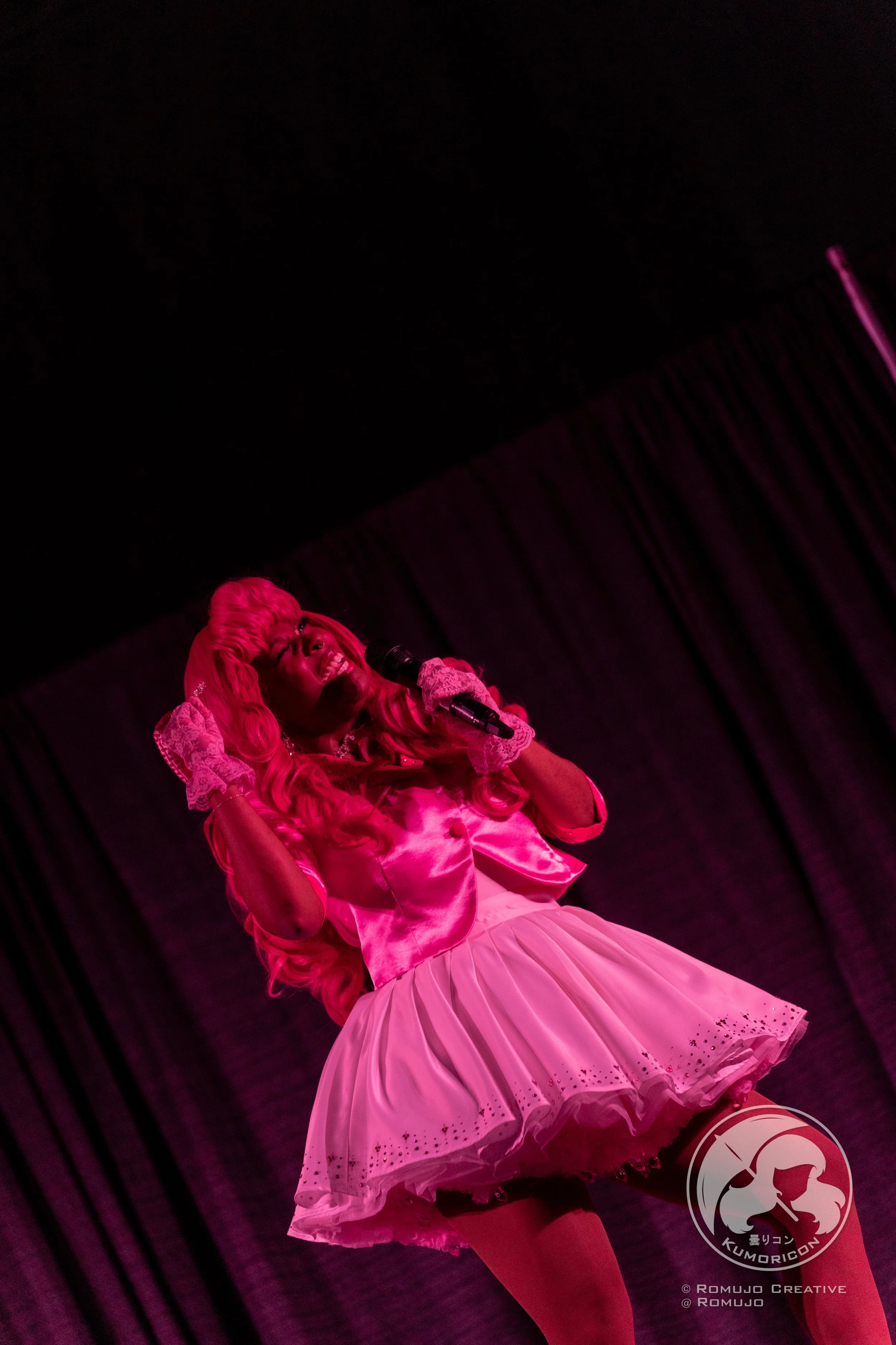 A woman dressed in a pink satin costume with a frilly pink tutu skirt, holding a microphone, performing on a stage with black curtains, lit in pink lighting.