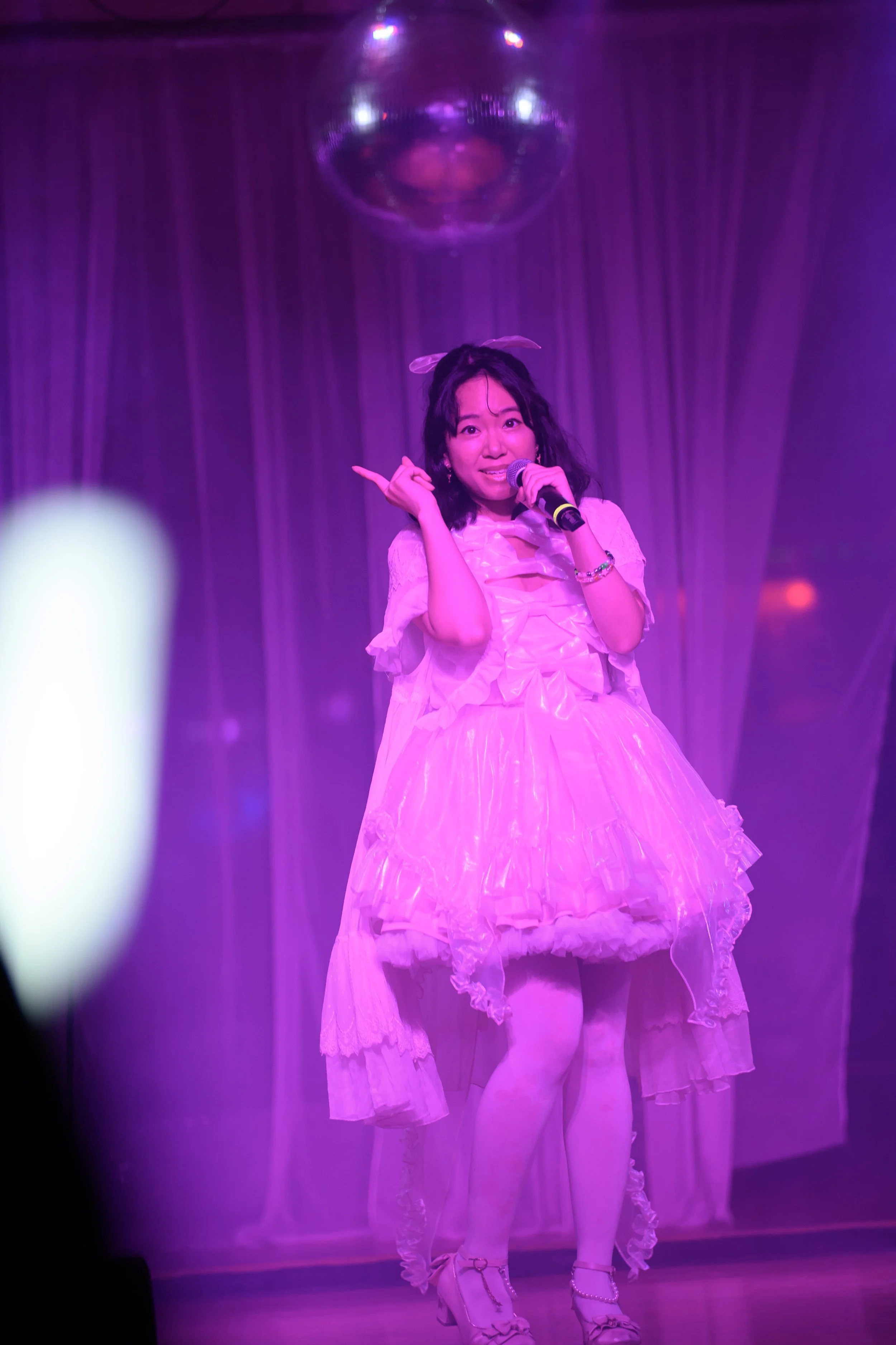 A woman dressed in a pink, frilly dress with a petticoat, singing or performing on stage with a microphone under purple stage lighting, with a disco ball overhead and curtains in the background.