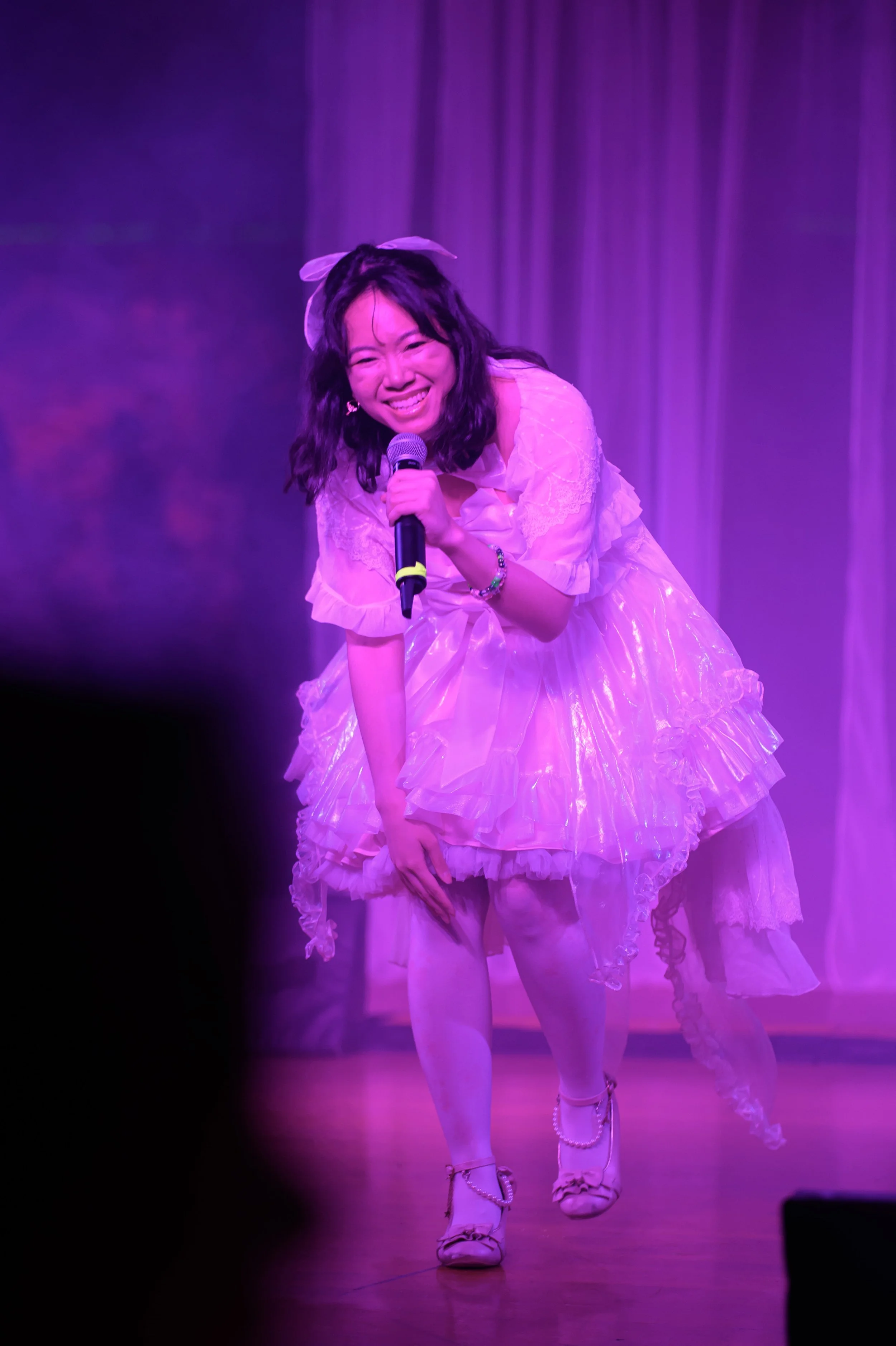 A woman in a pink dress with ruffles holding a microphone, smiling and bowing on stage with purple lighting and curtains in the background.