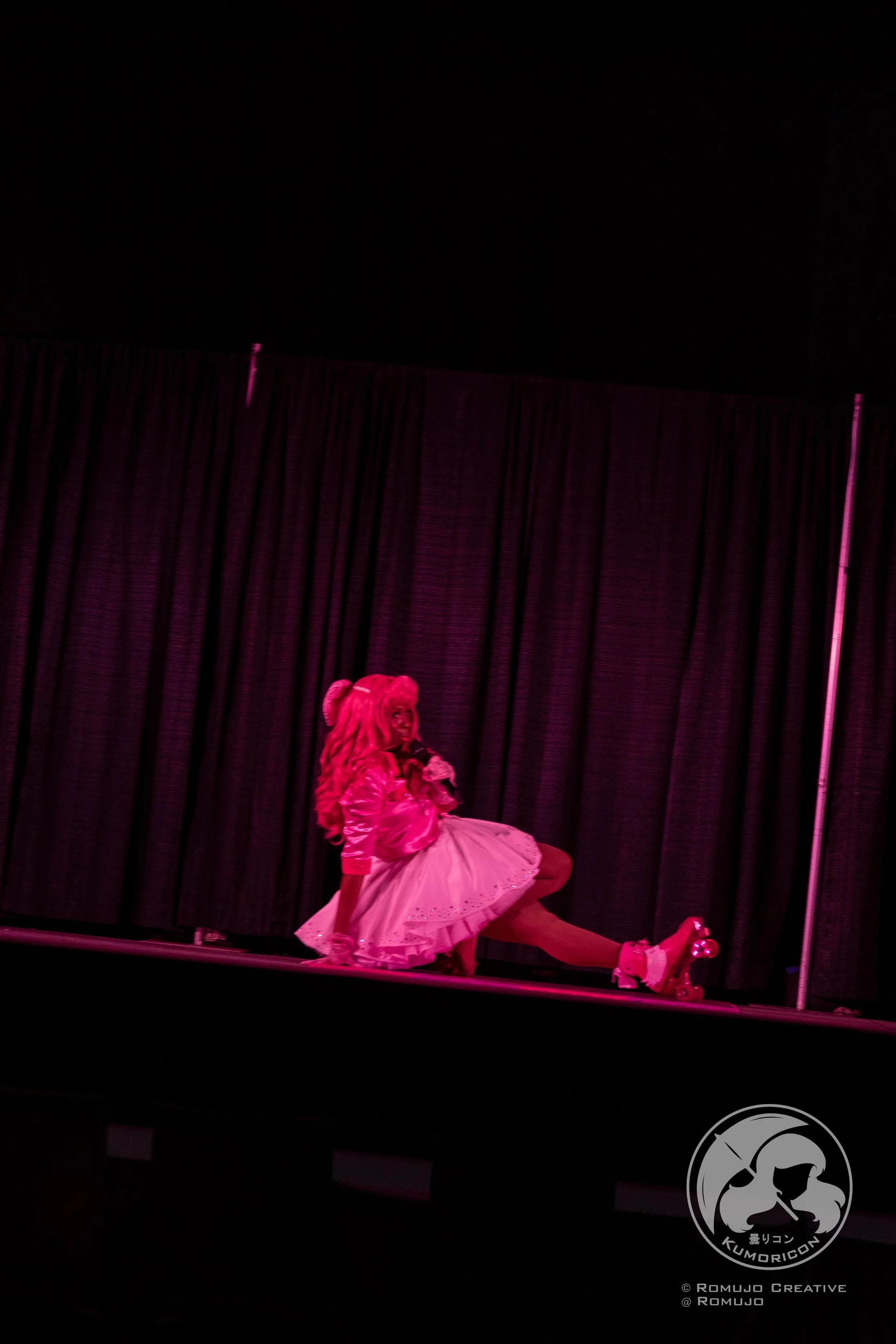 Person in pink dress on roller skates performing on stage with black curtains in the background.
