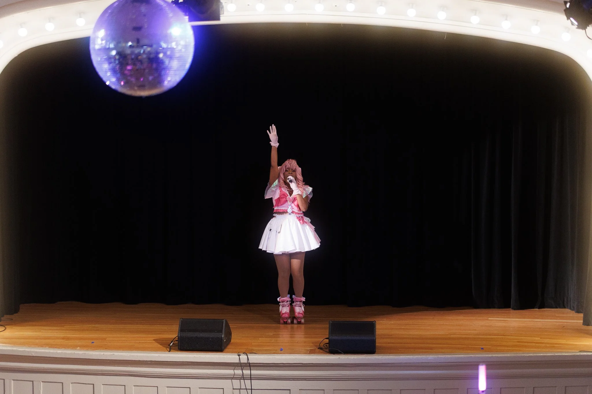 Performer on stage in pink and white costume, roller skates, singing into microphone, with disco ball overhead.