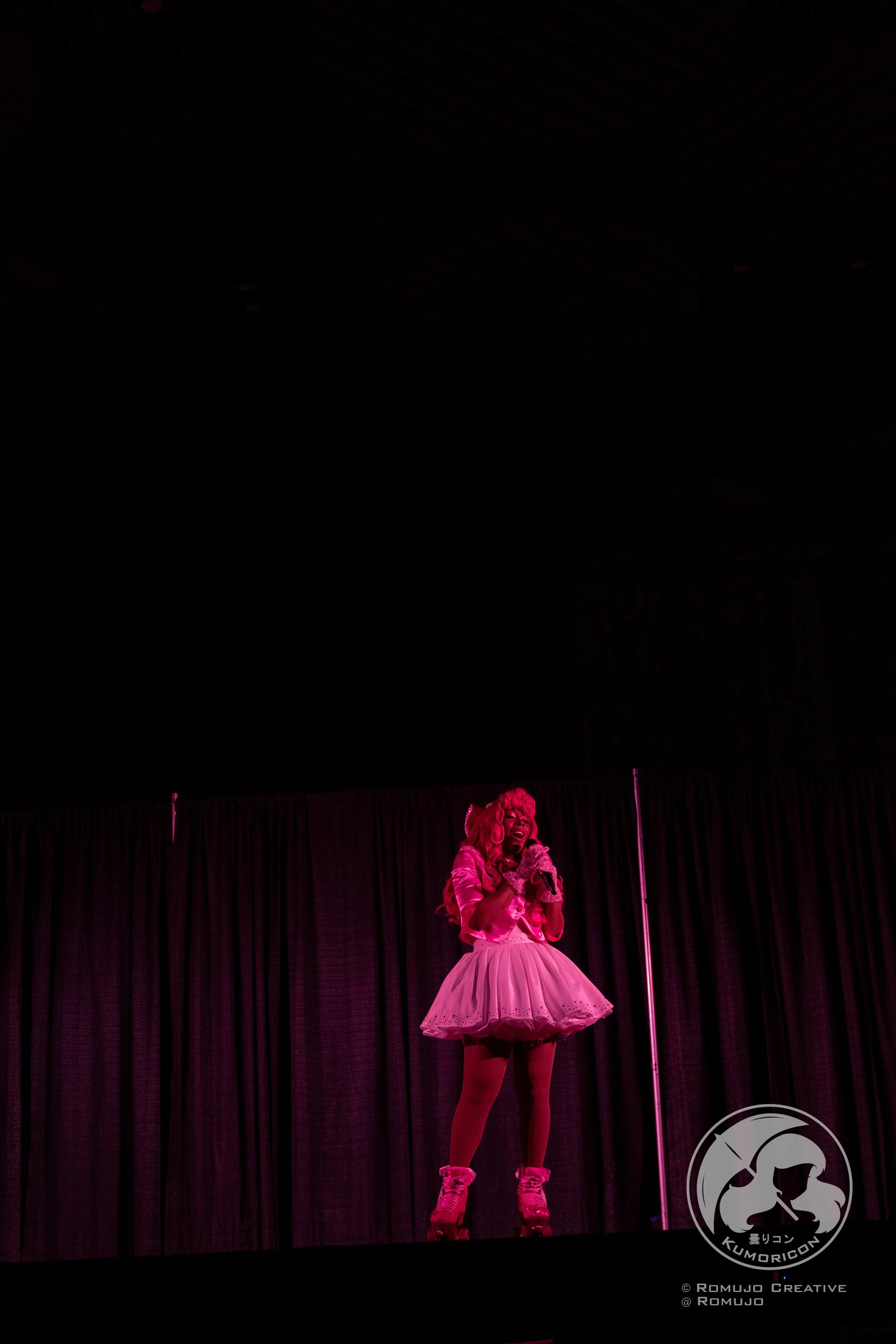 Performer on stage dressed in a pink costume with a tutu skirt, wearing roller skates, singing into a microphone, with a dark curtain backdrop.