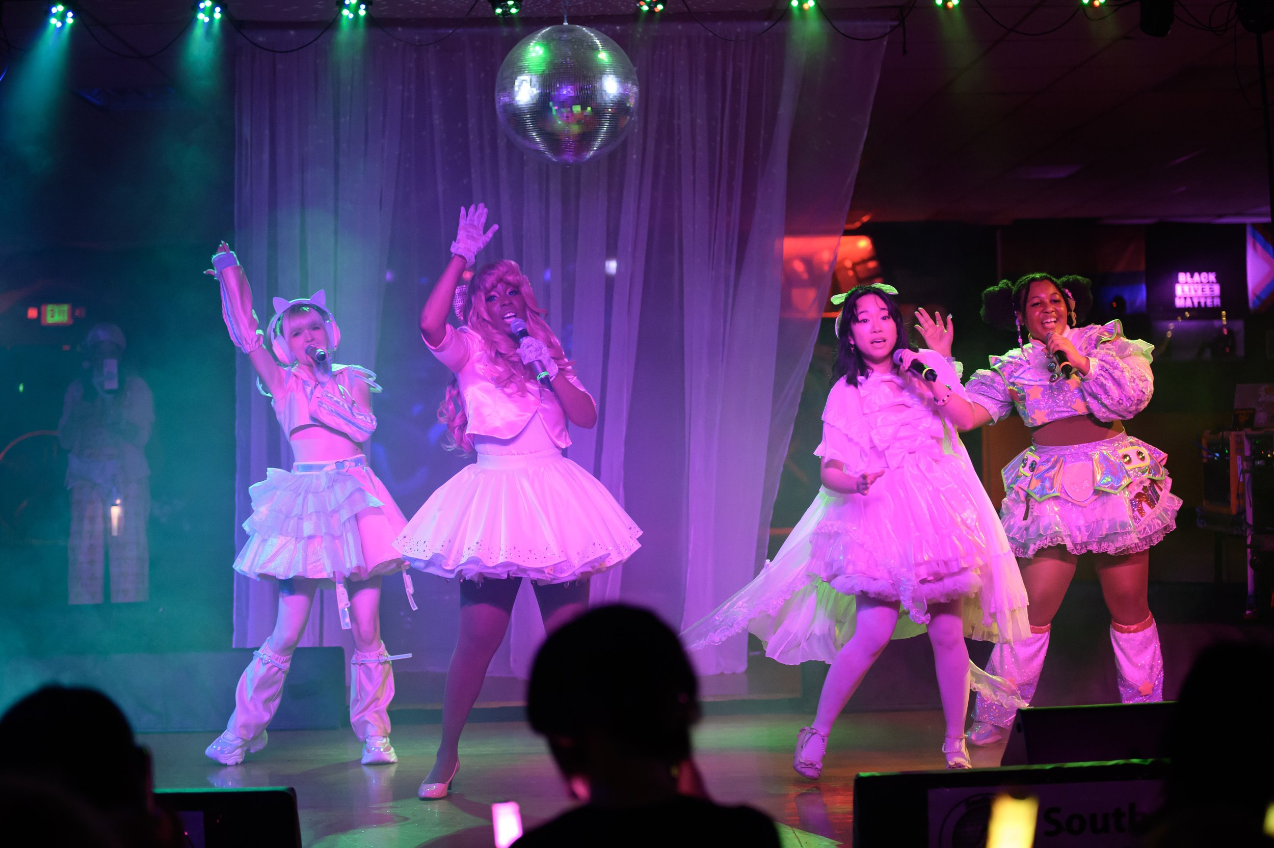 Four women in colorful, whimsical costumes performing on stage under green and purple lighting with a disco ball overhead.