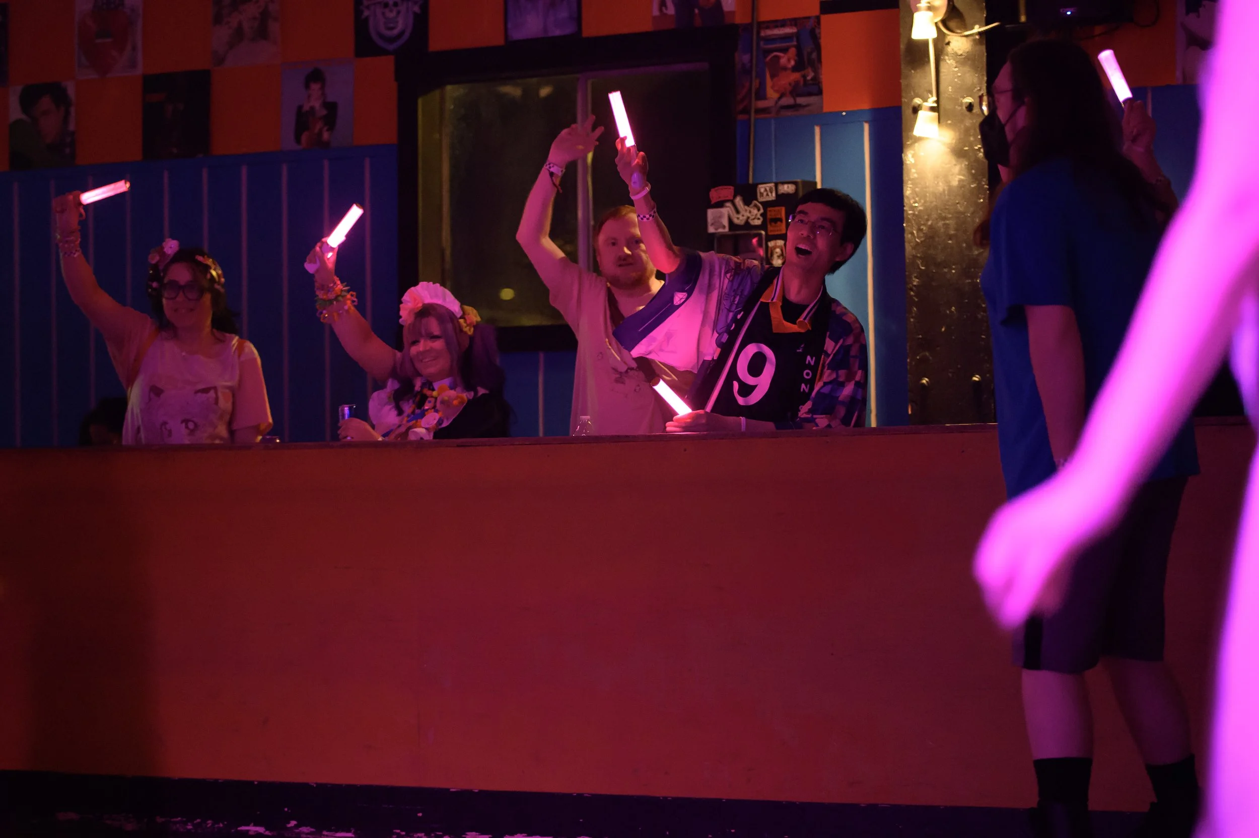 People celebrating in a dimly lit setting, holding pink glow sticks, with some dressed in colorful or floral accessories, in a vibrant, colorful environment.