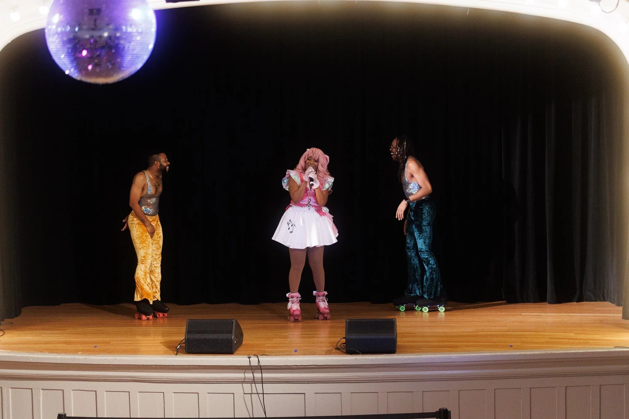 Three performers on stage roller skating, with a woman in center singing into a microphone. The woman has pink hair and is dressed in a white skirt with musical notes, and the men are wearing colorful, shiny tank tops and velvet pants. A disco ball h
