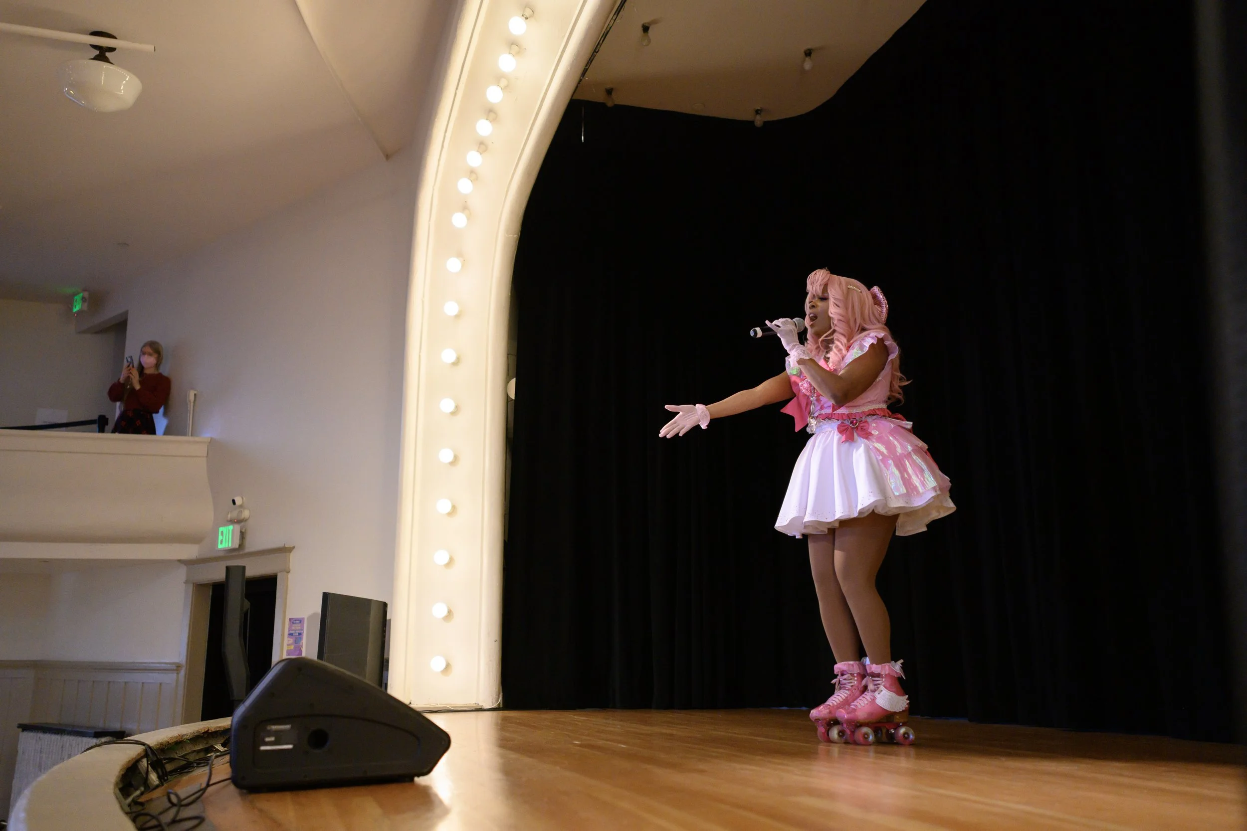 A performer dressed in pink and white roller skates, a pink wig, and a pink and white dress singing into a microphone on stage with black curtains. An audience member in the background takes a photo.