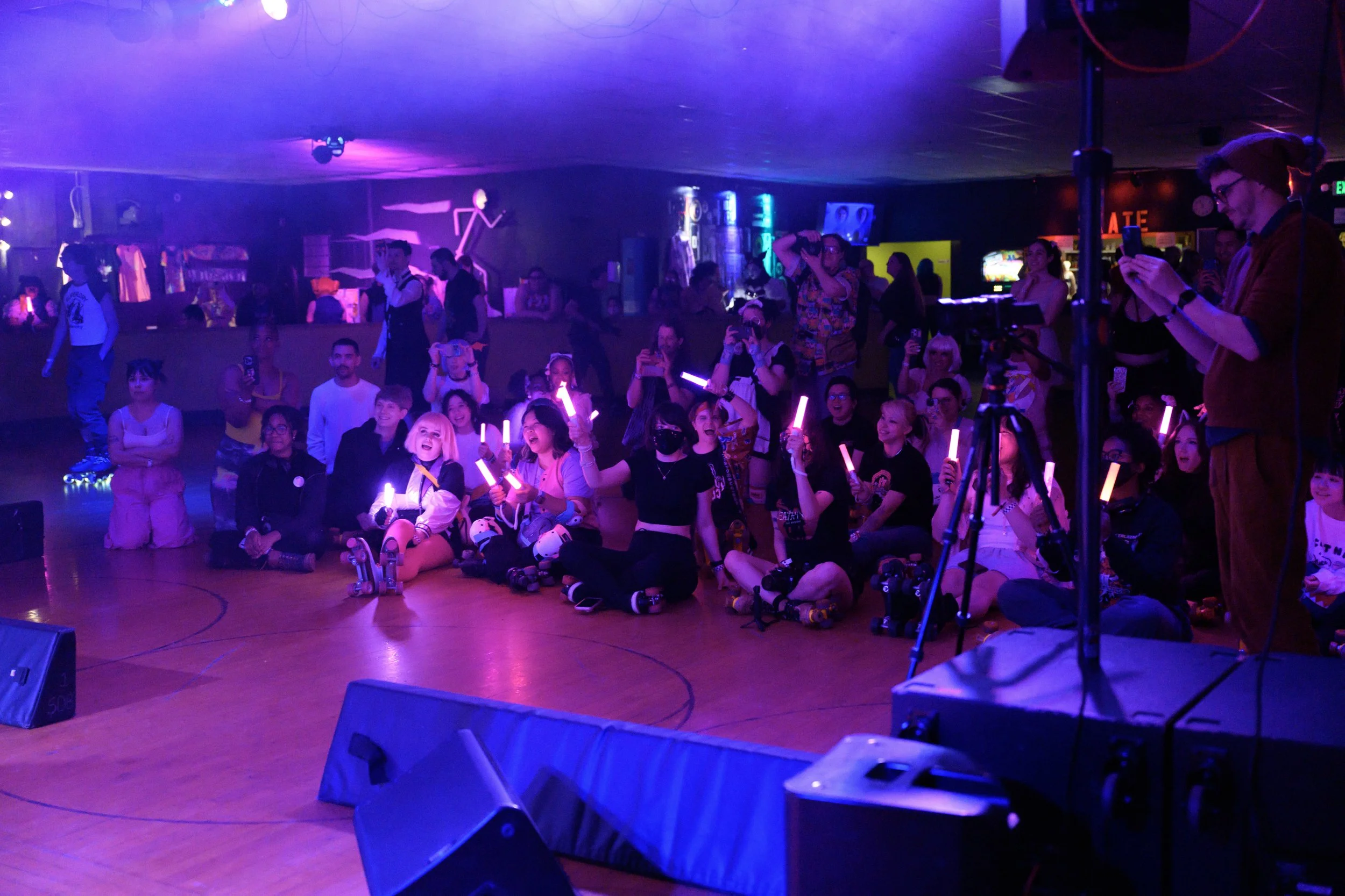 Group of children and teenagers sitting on the floor at a roller skating event, holding glowing pink and orange light sticks, with some people taking photos and videos, in a dimly lit venue with purple and blue lighting.