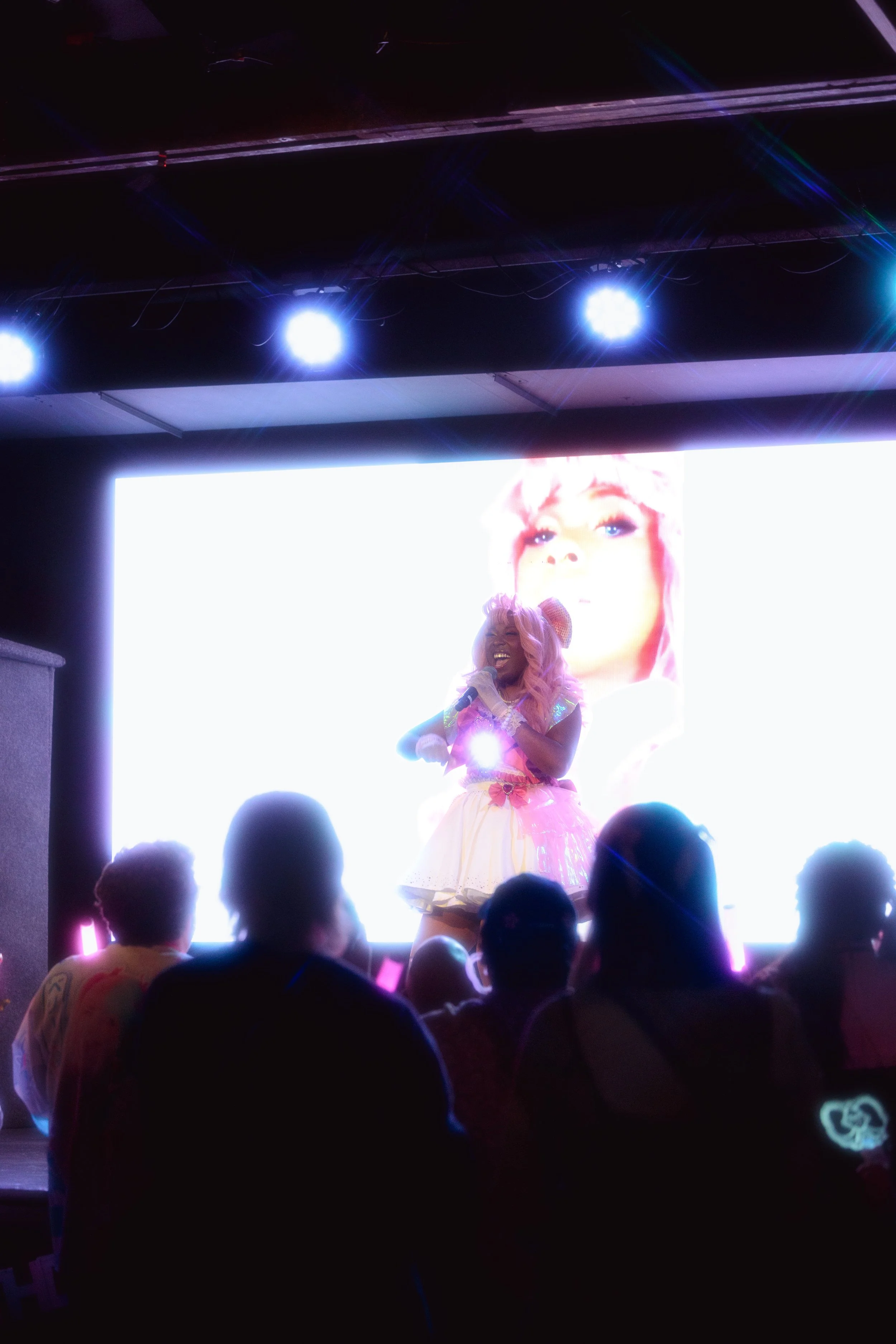 Performer on stage dressed in a colorful, whimsical outfit with pink hair, holding a microphone, in front of a large bright screen with a close-up image of a woman's face. Audience members are seated facing the stage.