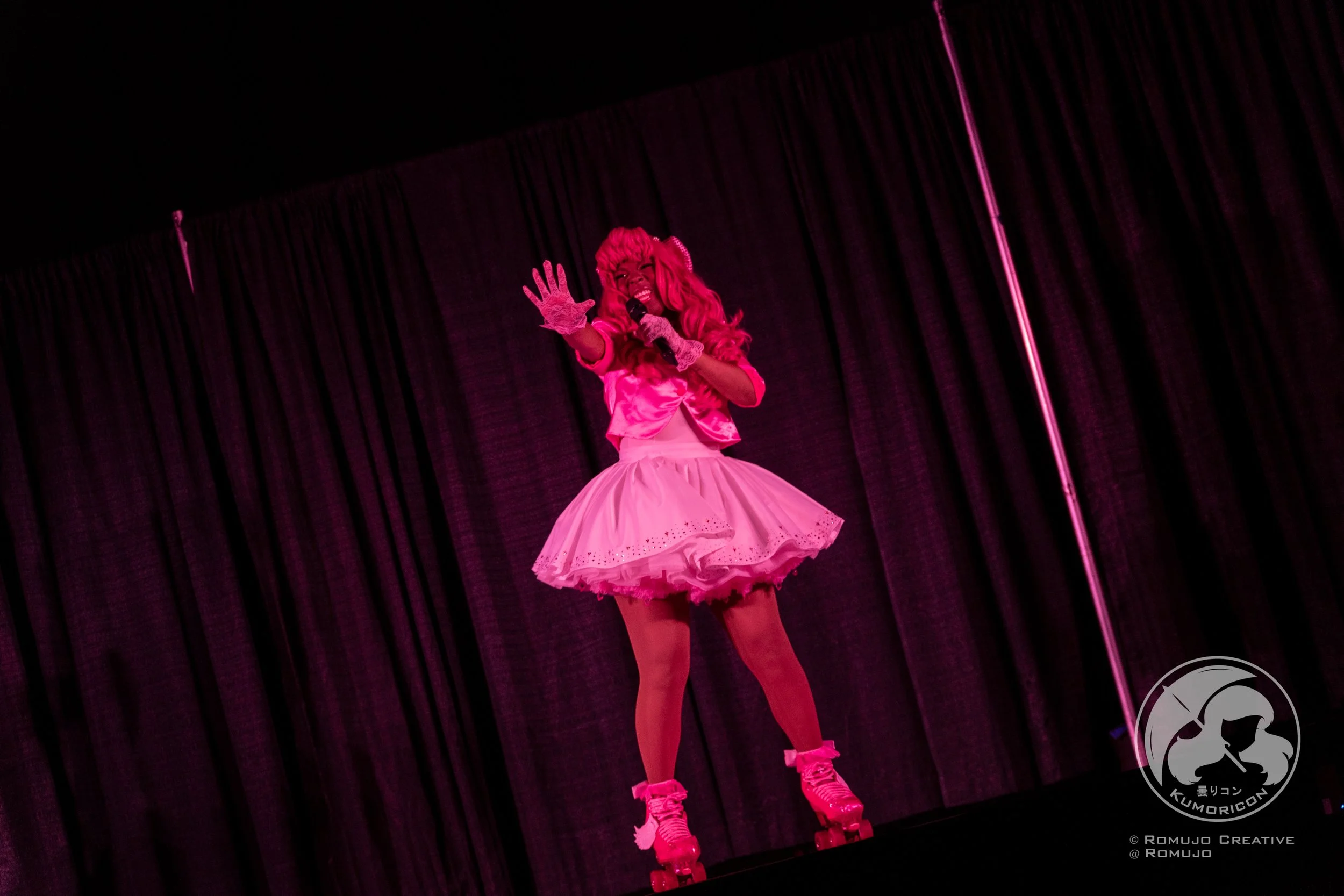 A performer dressed in a pink outfit, including a wig, gloves, and roller skates, singing on stage with a microphone, in front of a black curtain.