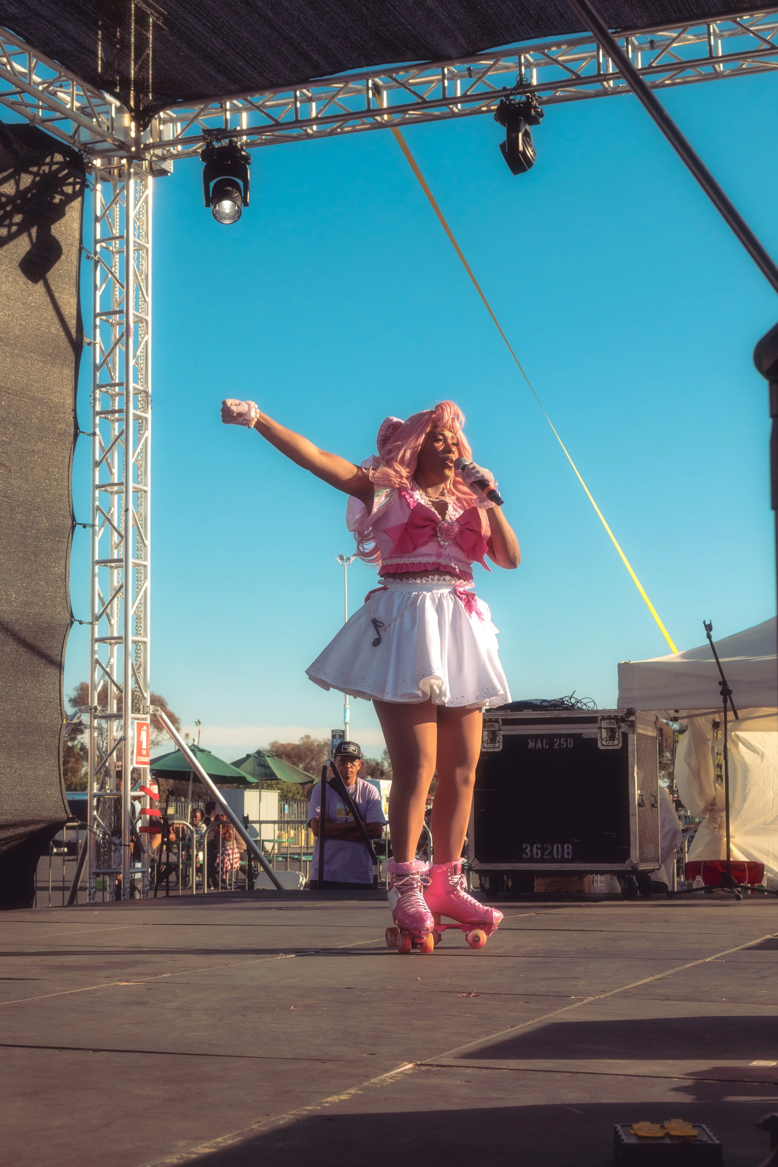 A woman dressed in pink and white roller skates, a pink and white outfit, and pink hair, performing on stage with a microphone under a blue sky.