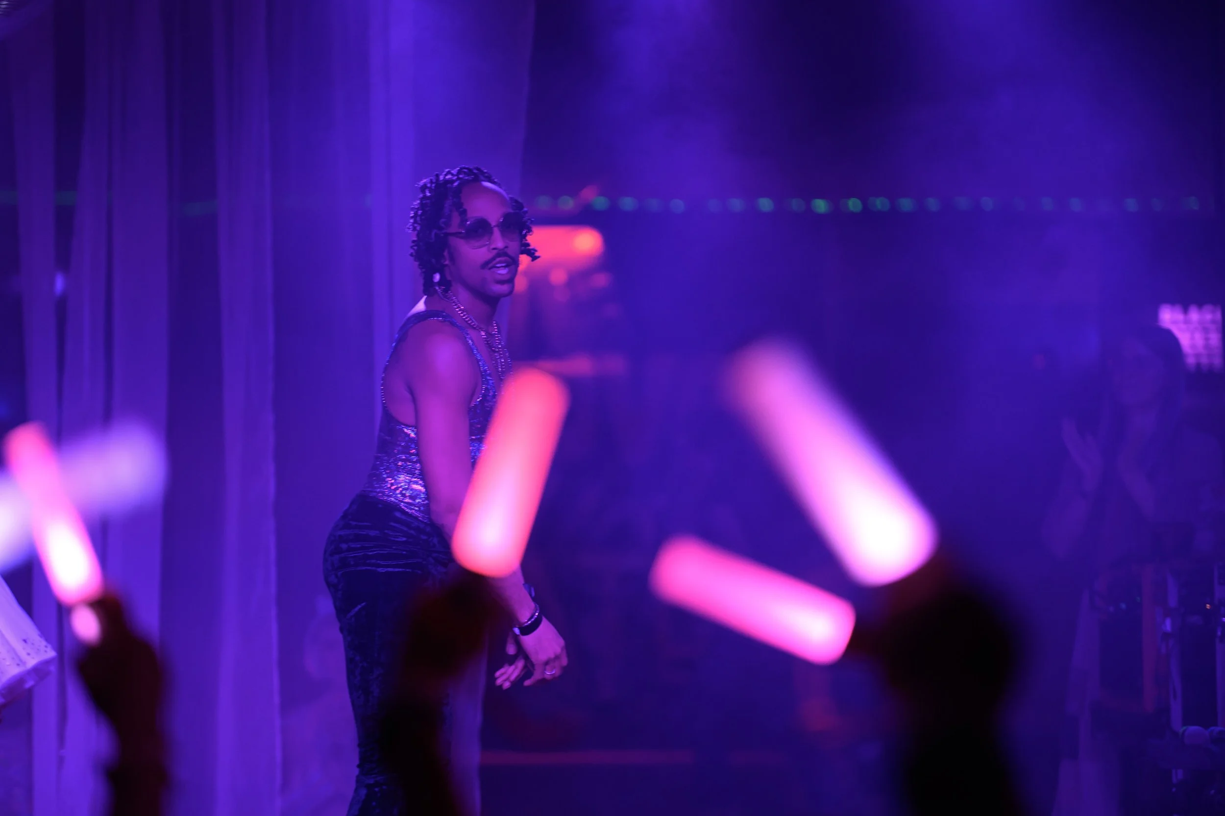 A person with dreadlocks and sunglasses standing in a dimly lit space illuminated by purple and pink lights, holding glow sticks.