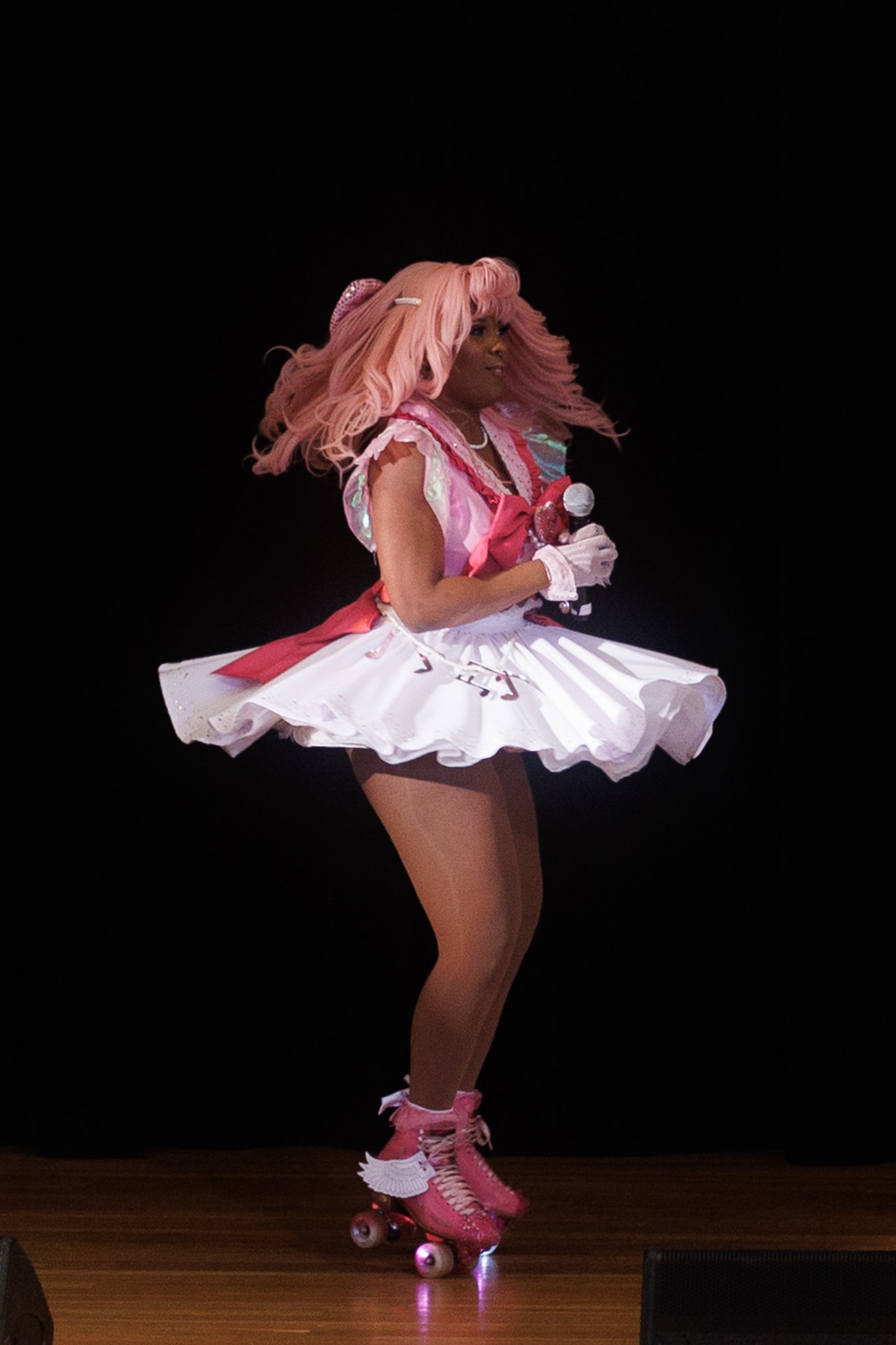 Person wearing a pink wig, pink and white outfit, pink roller skates, and holding a microphone on a stage.