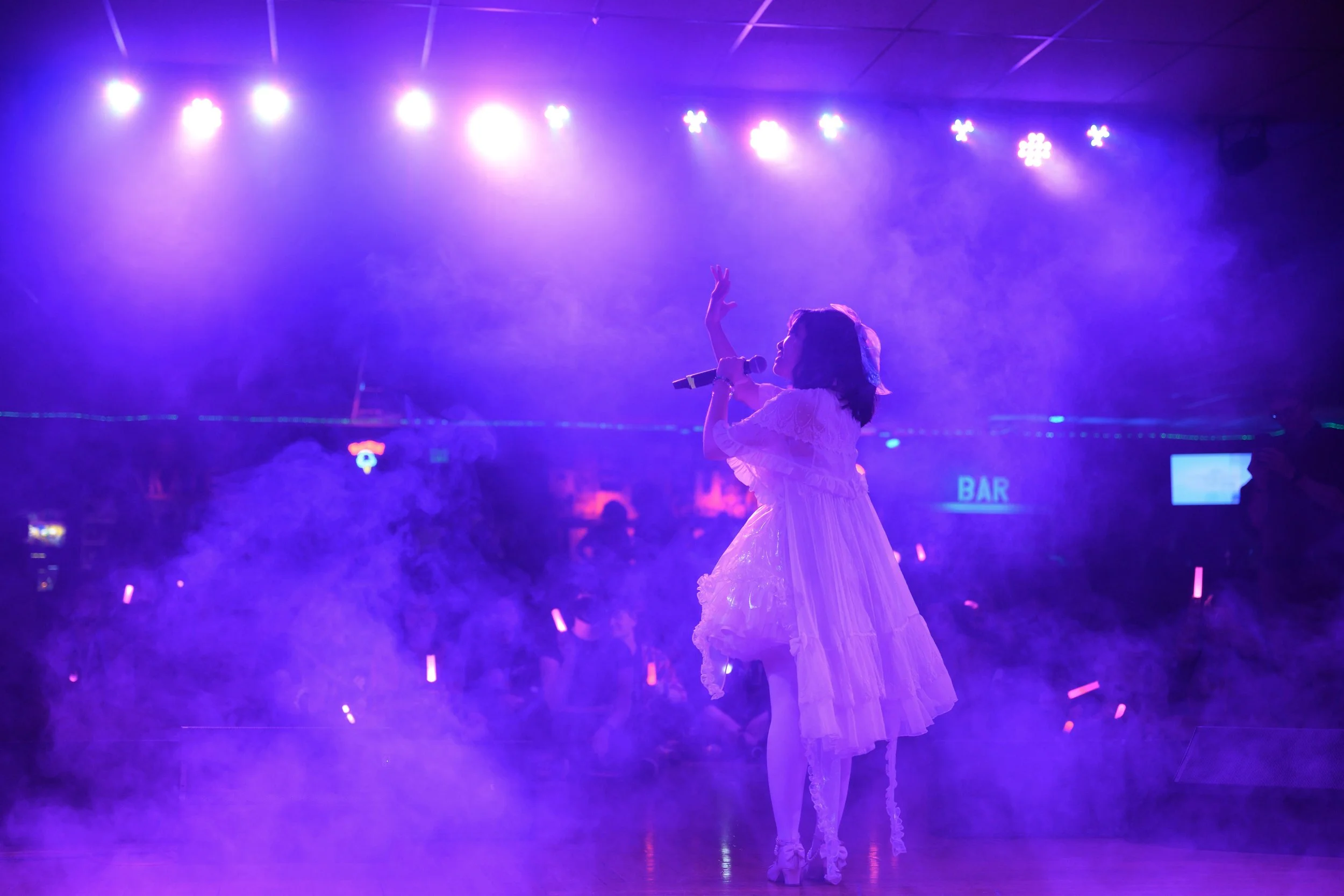 A woman singing on stage under purple lights with fog, wearing a white dress and holding a microphone.