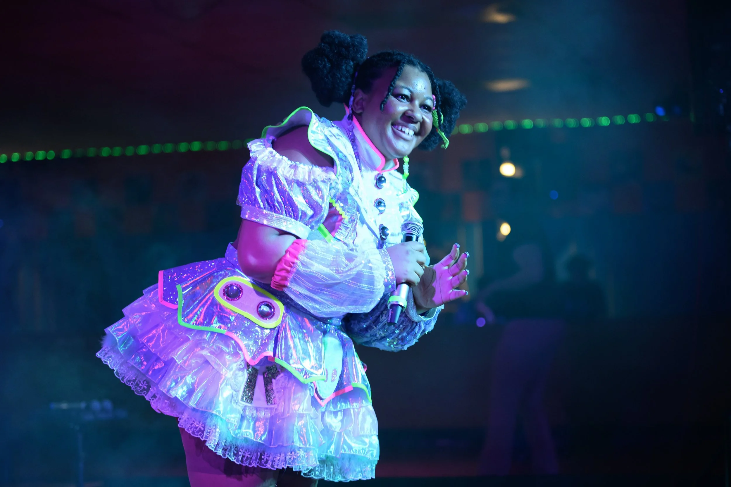A woman in a colorful, shiny costume with puffy sleeves and ruffles, smiling and holding a microphone on a stage with vibrant lighting.