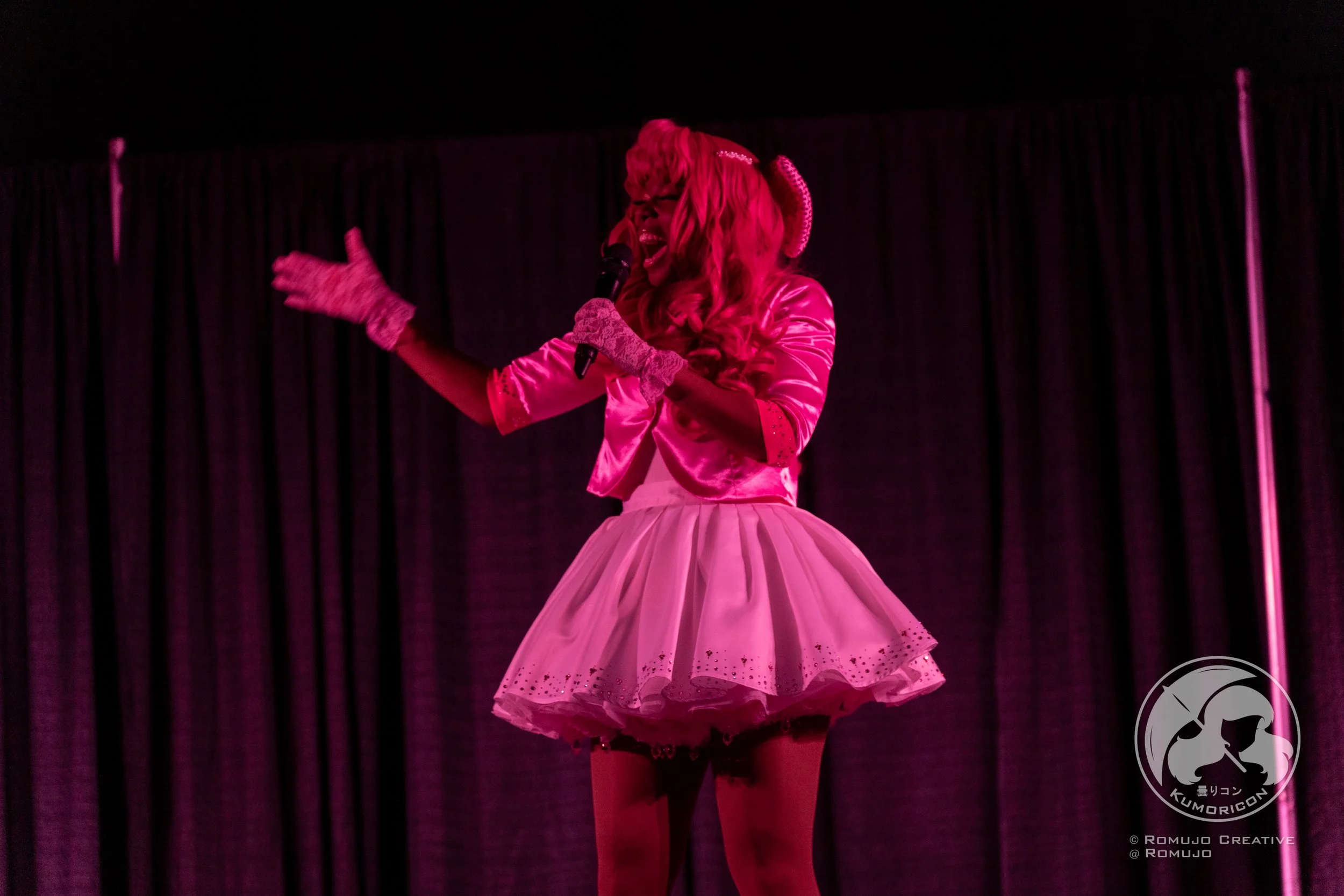 Performer on stage wearing a pink satin jacket and a fluffy pink skirt, singing into a microphone with a black curtain backdrop.