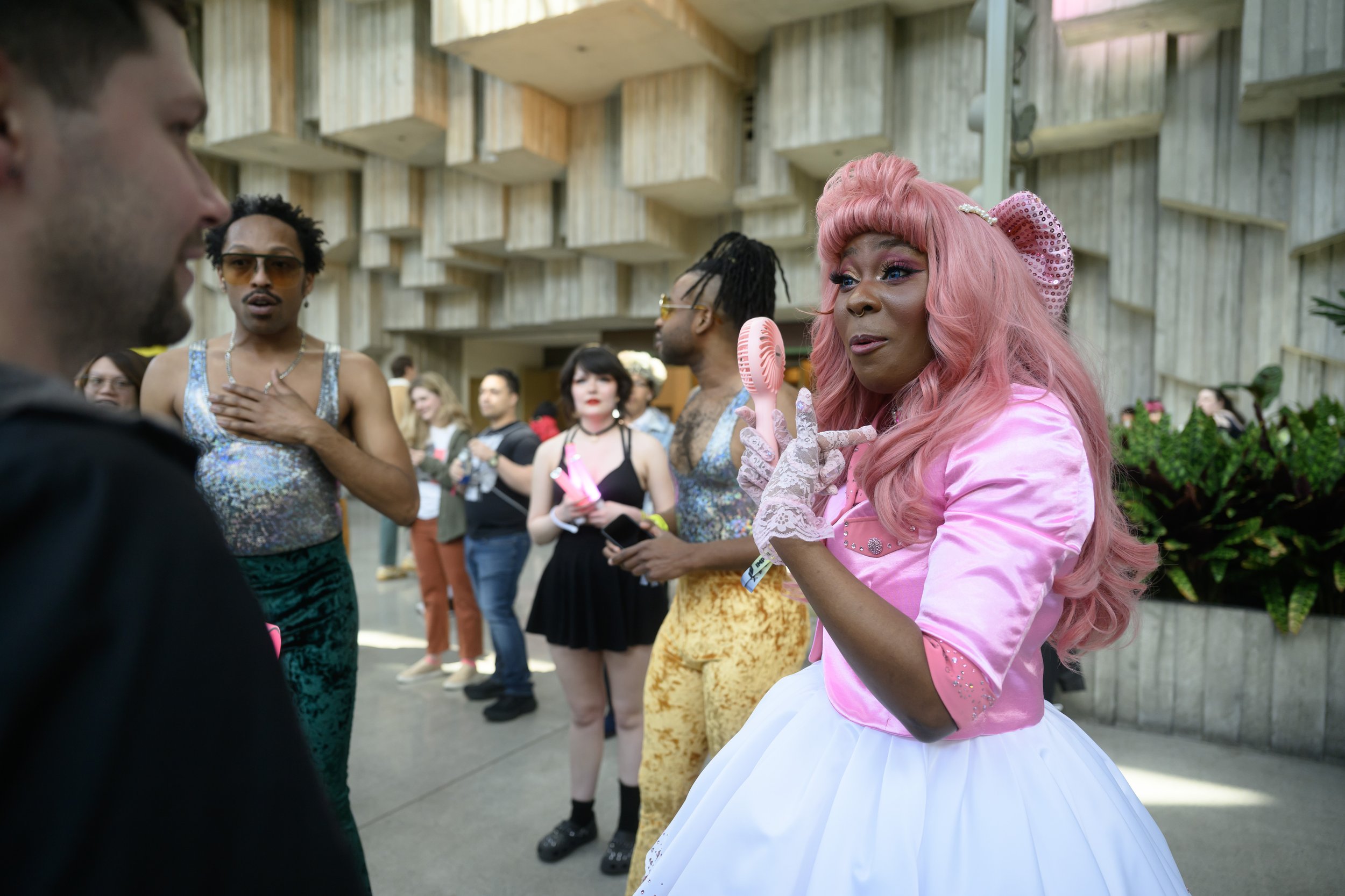 A woman in a pink costume with a wig and gloves is holding a pink hairbrush and talking to a man at a social event. Several people are in the background, dressed in colorful and eclectic outfits.