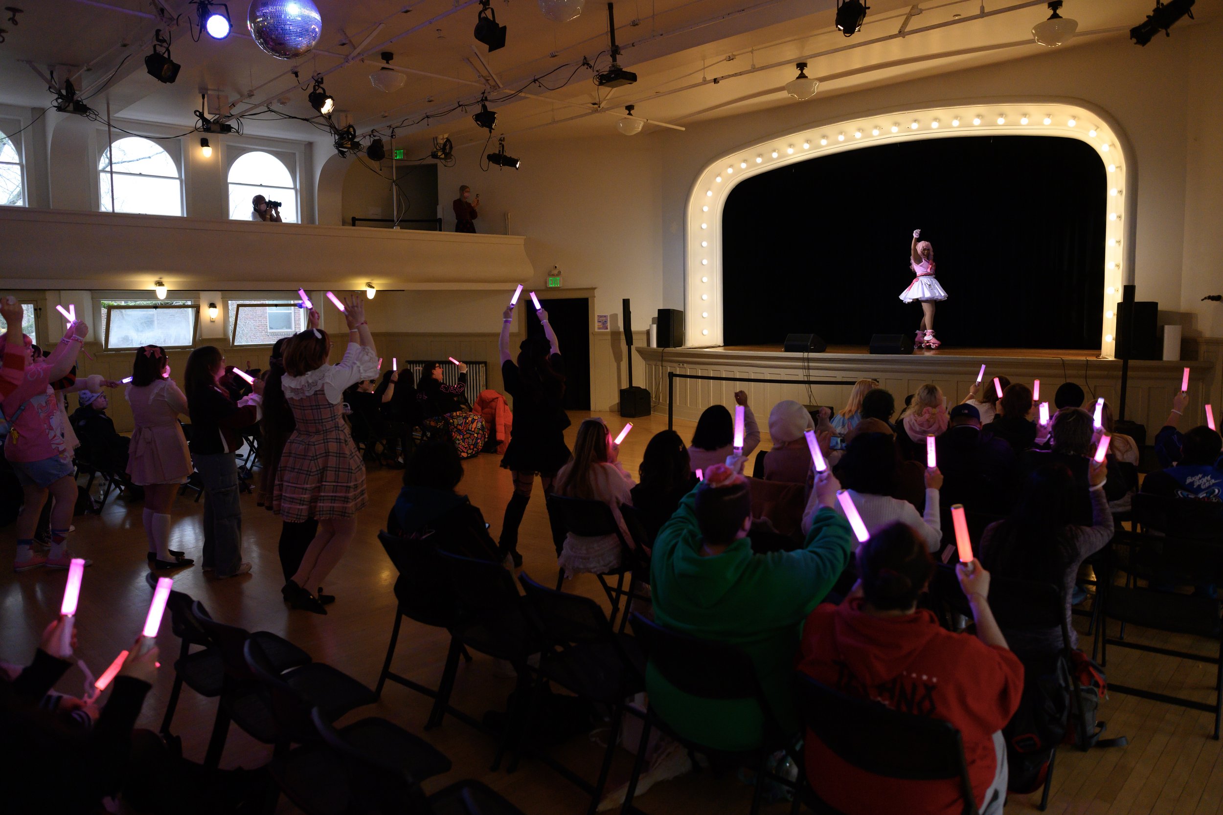 A girl dressed as a ballerina performing on stage in front of an audience, holding a pink rose. The audience, mostly children, seated and standing, are using pink glow sticks, in a dimly lit theater with stage lights and a large black curtain.