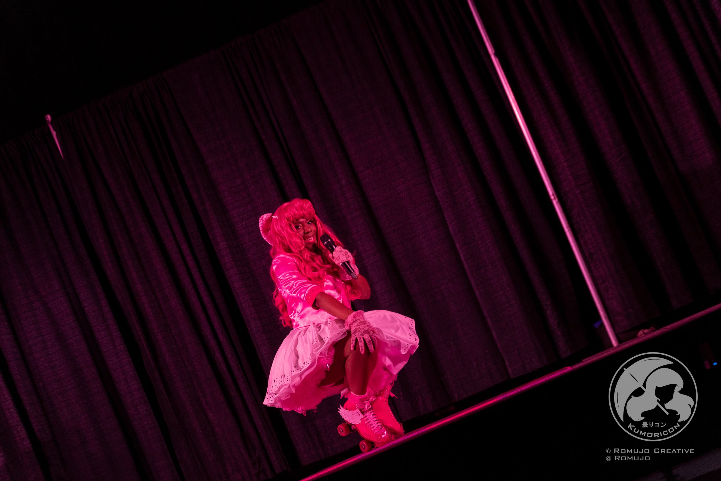 A person in a pink wig and matching costume crouching on roller skates, singing into a microphone with a dark curtain backdrop.