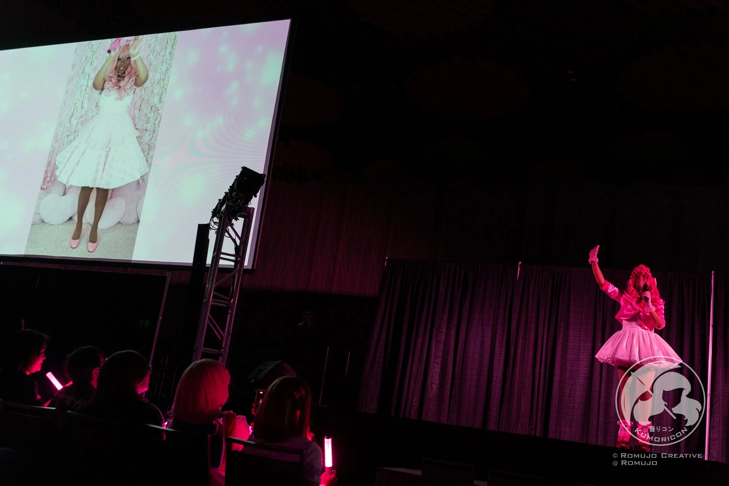 A stage with a performer dressed in pink, wearing a curly pink wig, pink gloves, and a pink tutu, speaking into a microphone. Audience members are seated, some holding pink glow sticks. A large screen behind displays a woman in a white dress with pin