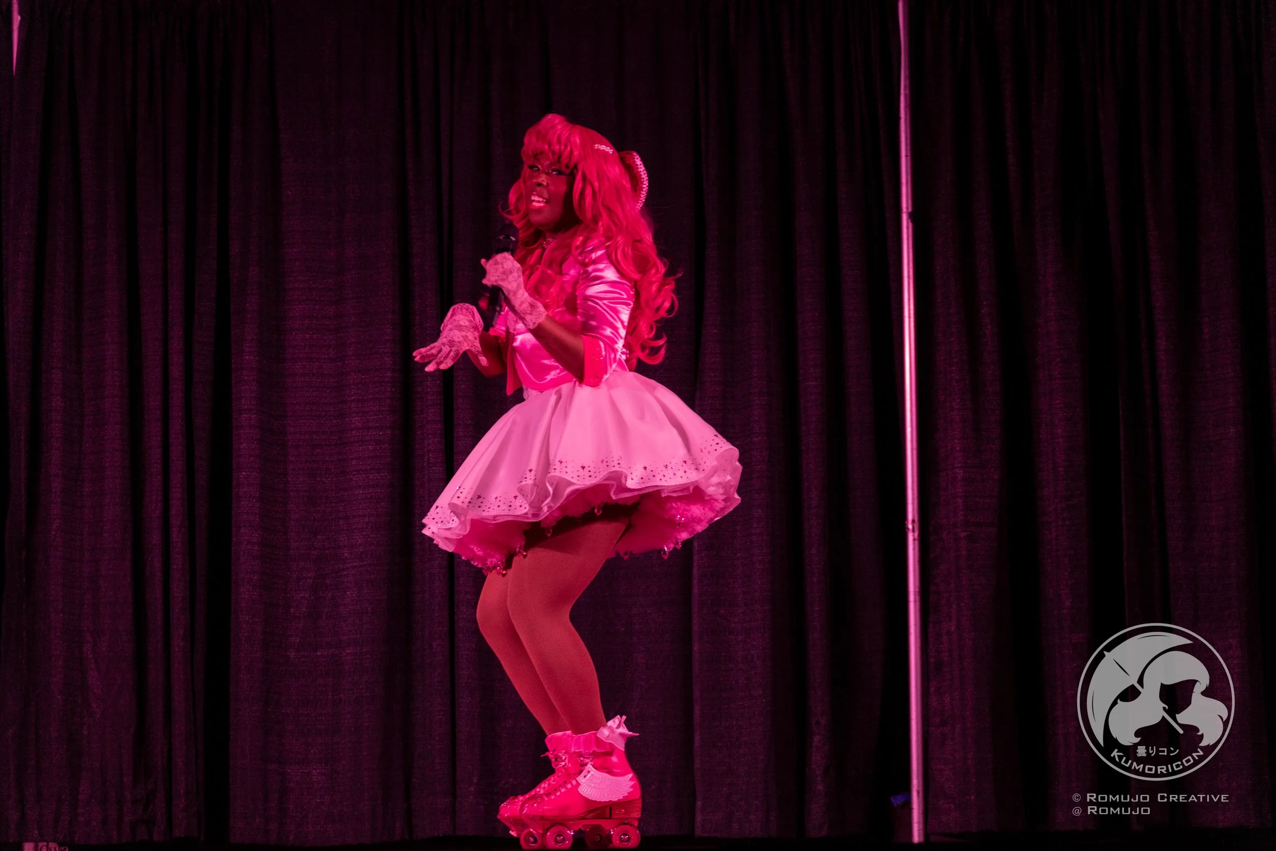 Performer with pink hair in a pink and white dress on stage, roller skating under pink lighting with black curtains in the background.
